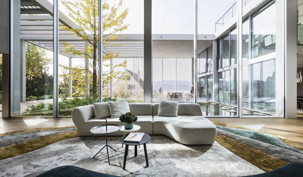Double-height living room with floor-to-ceiling windows. Curved off-white sofa, organic-shaped rug in earth/green tones. Outside view of the garden and water basin.
