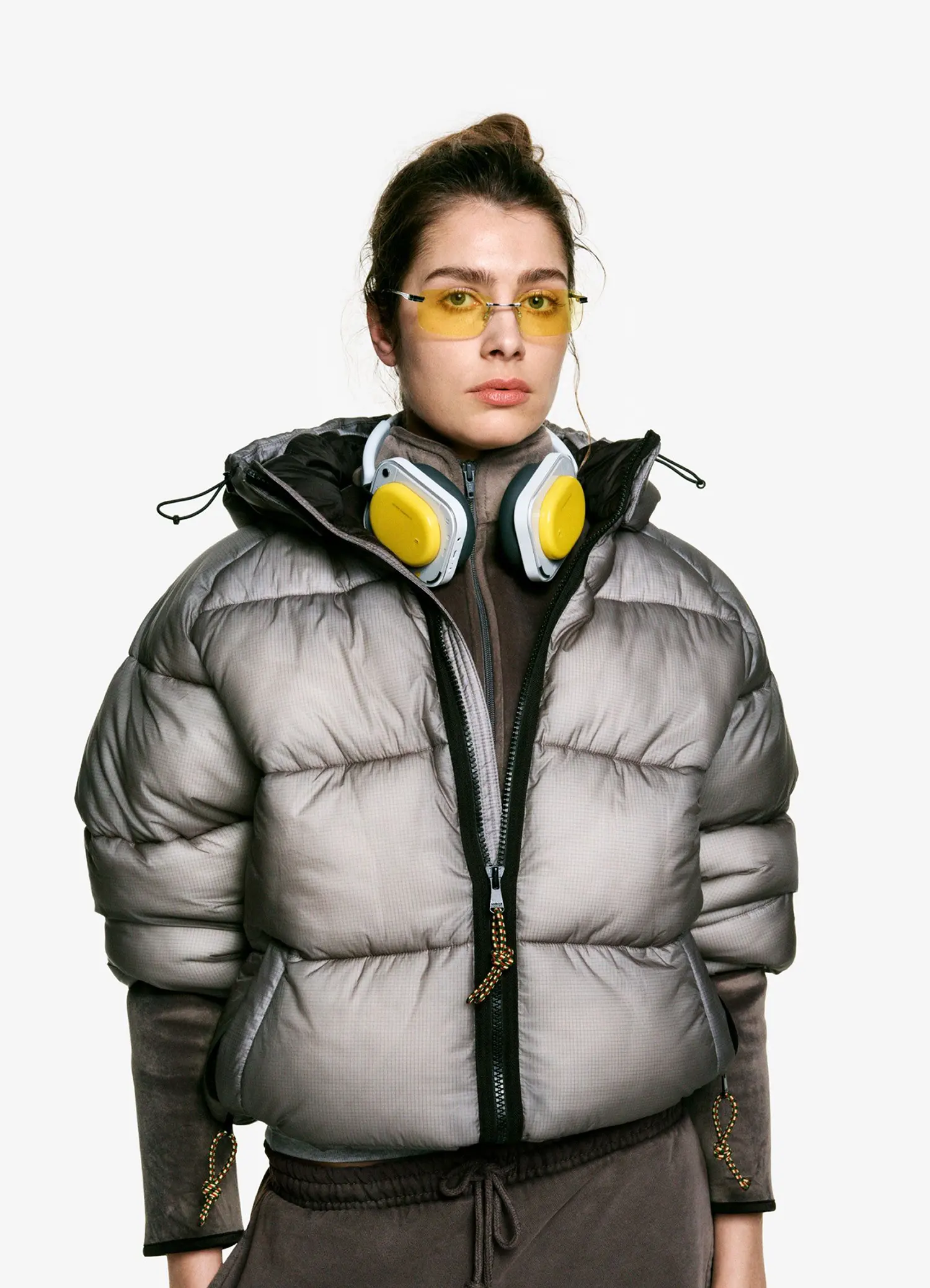 Woman wearing yellow Nothing Headphone (a) around her neck with a grey puffer jacket.
