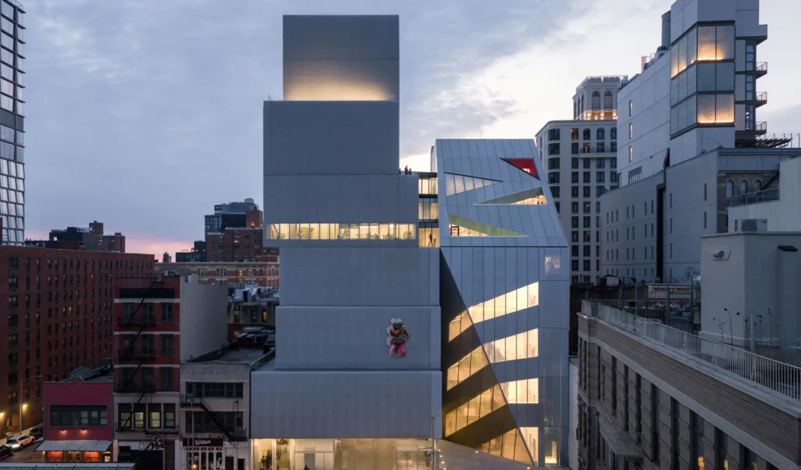 Exterior of OMA’s prismatic New Museum extension at dusk with glowing interior circulation.