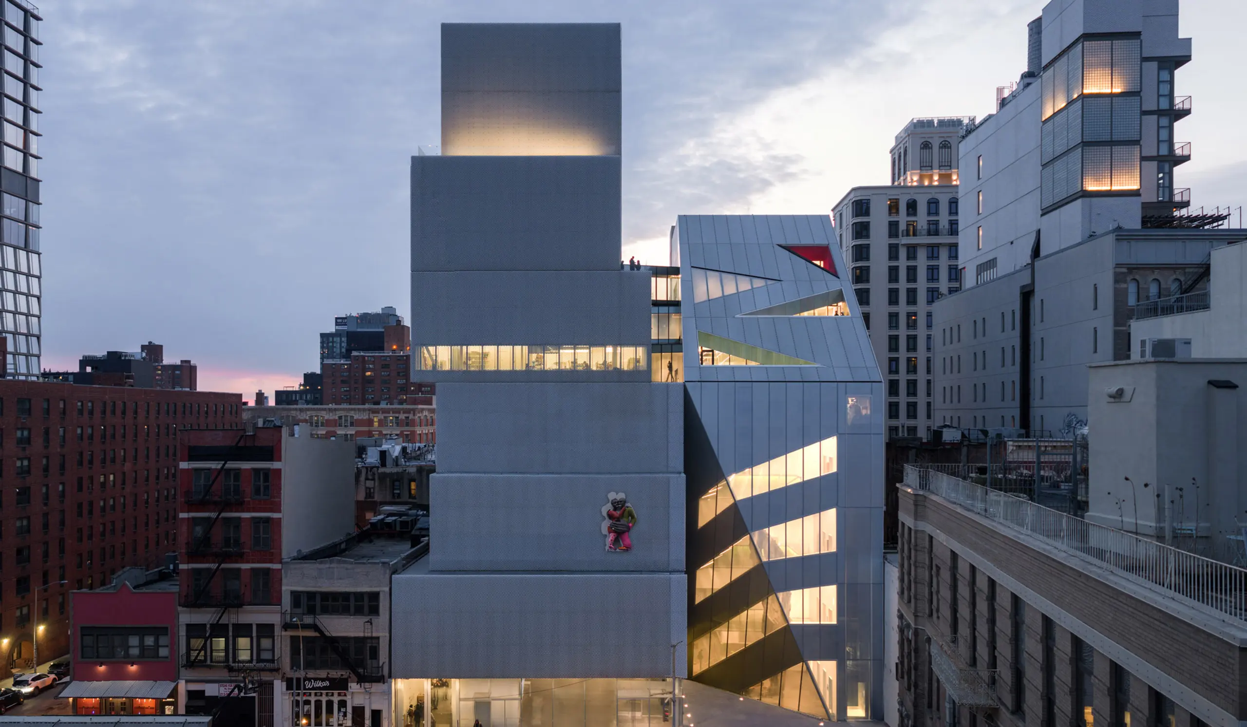 Exterior of OMA’s prismatic New Museum extension at dusk with glowing interior circulation.