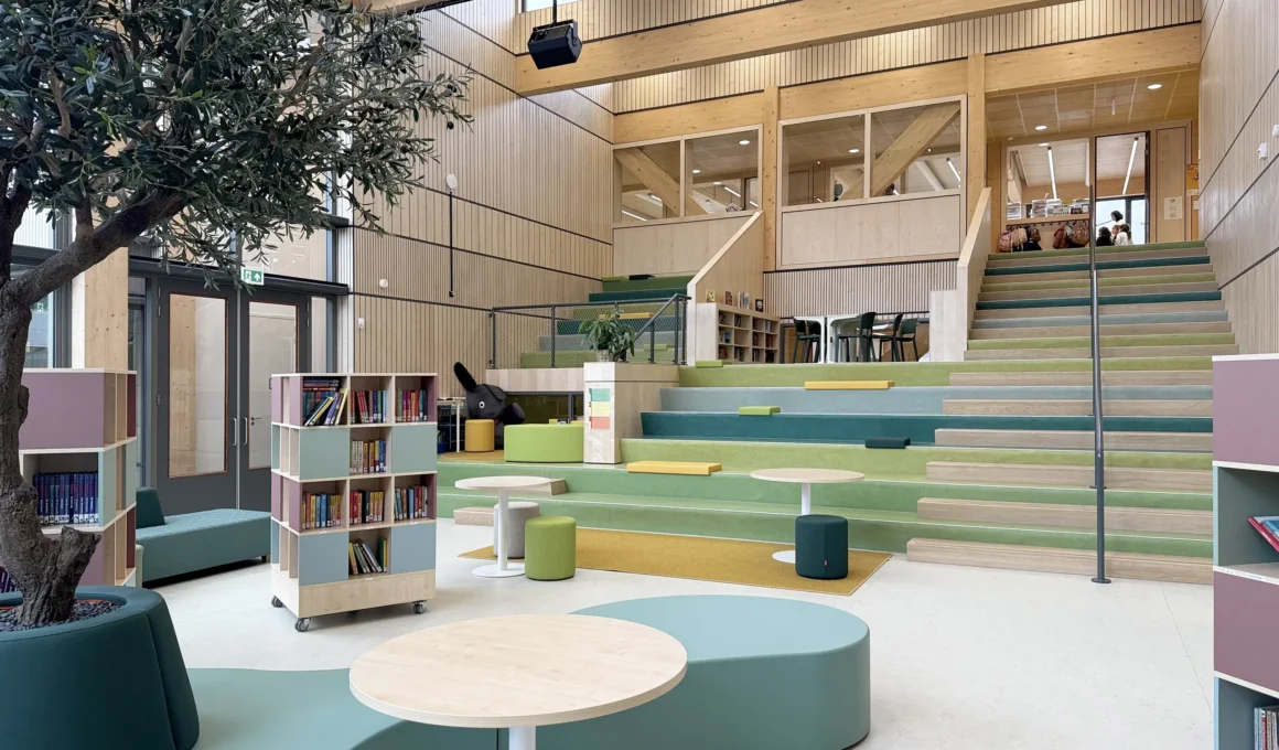 A sunlit central auditorium with tiered, green-padded seating, a library nook, and wooden architecture at Wisperweide.