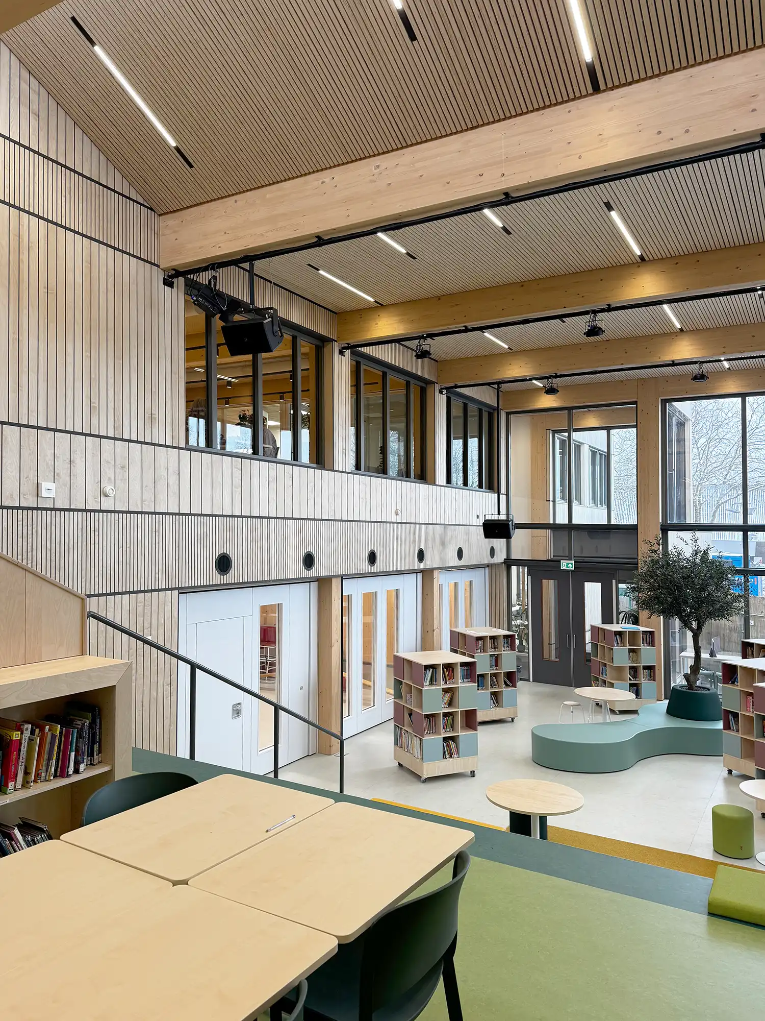 An elevated view of the high-ceilinged central auditorium, showcasing modular wooden architecture, a reading tree, and CLT beams.