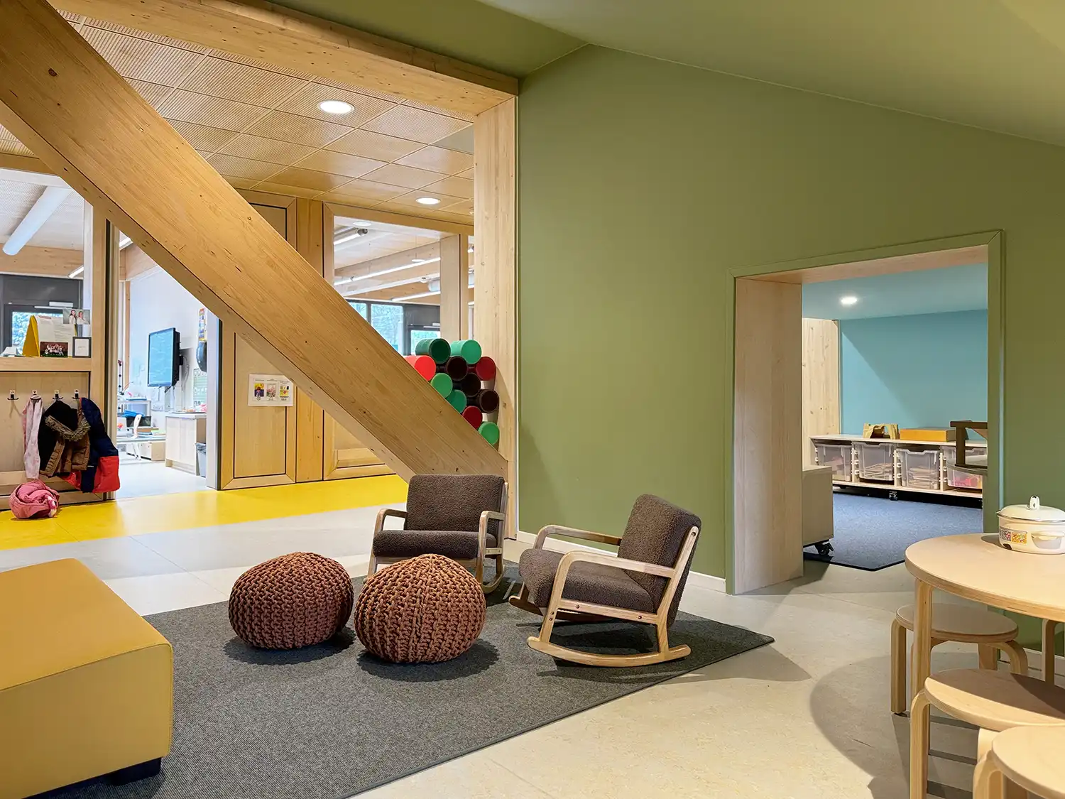 An angled perspective of a cozy reading nook with textured walls, woven ottomans, and a direct view into an active wooden classroom.