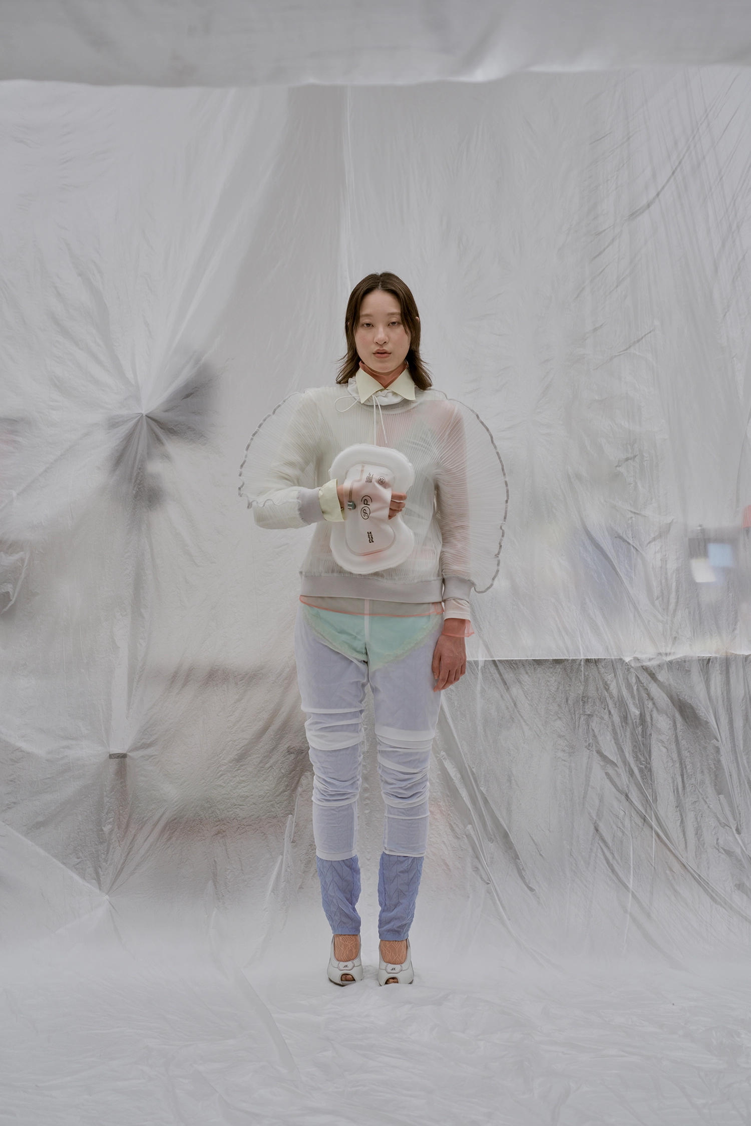 Full-body shot of a person wearing an avant-garde white outfit, holding the Pulse Pack at chest level against a translucent plastic backdrop.