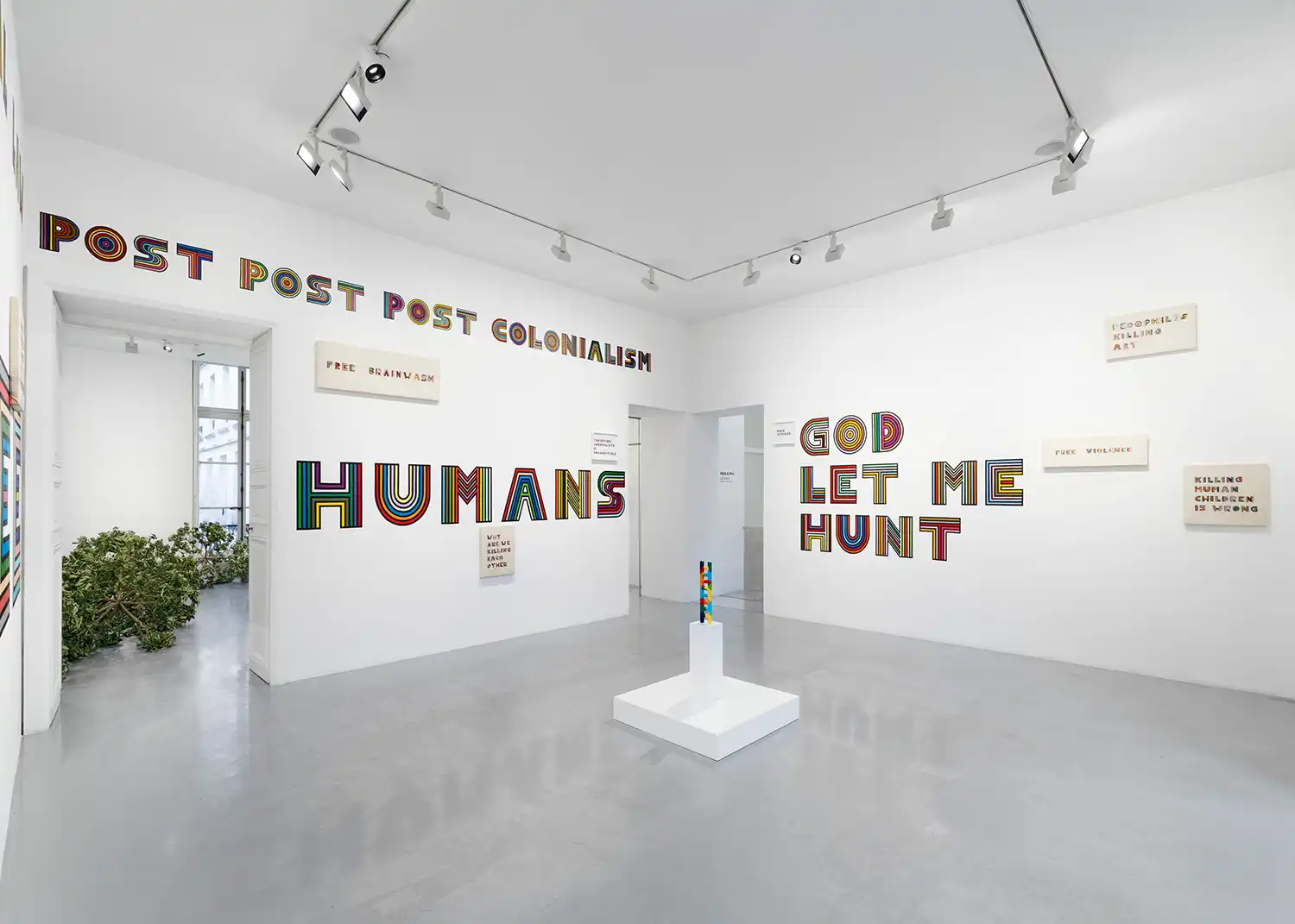 Perspective view of a white gallery room filled with large rainbow-colored embroidered text and a central pedestal.