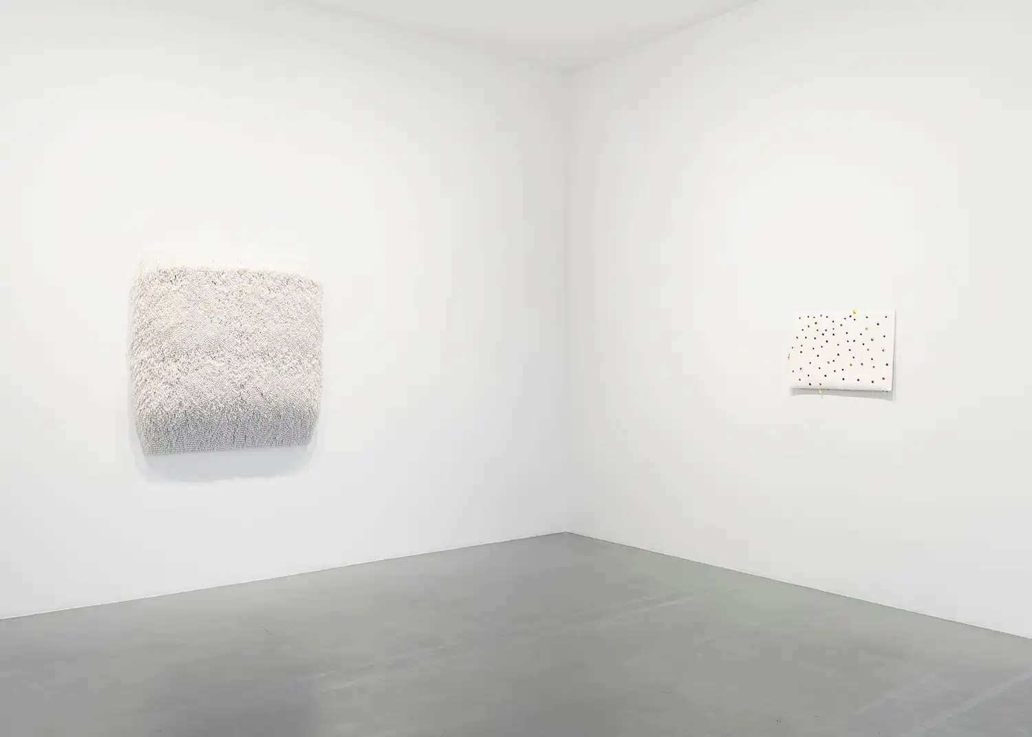 Two wall-mounted white sculptures: one a dense accumulation of pearls and the other a minimalist dotted canvas.