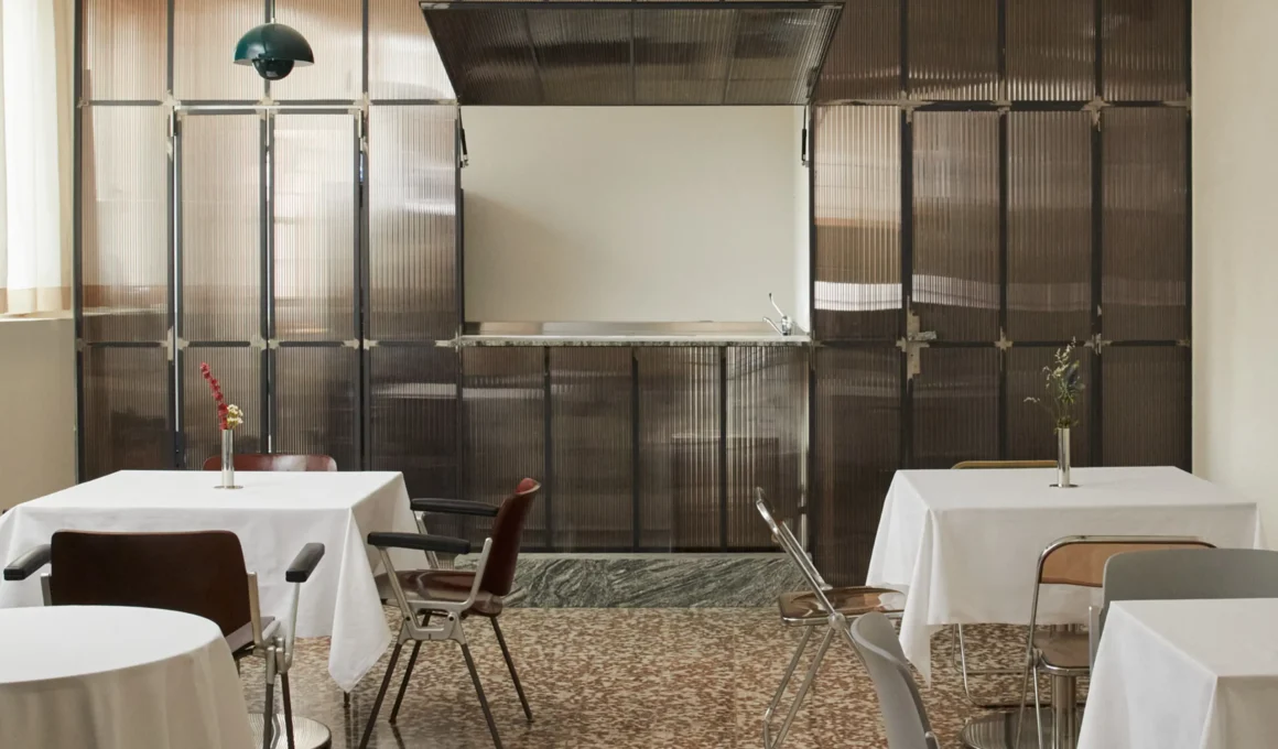 A spacious interior view of Sandì restaurant in Milan, showcasing a wall filter made of smoked polycarbonate panels and brushed steel frames separating the dining area from the kitchen, with white tablecloths on tables and a terrazzo floor.