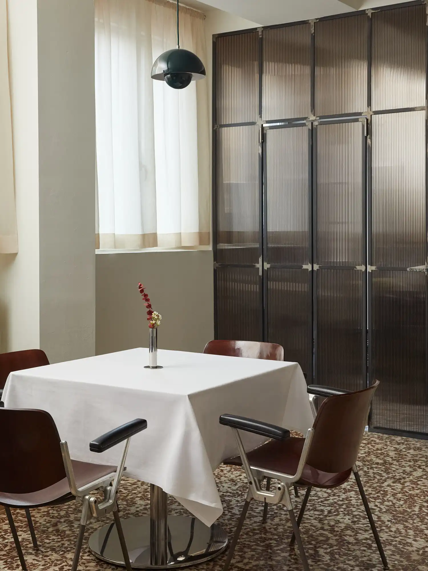 A close-up view of a dining table at Sandì restaurant, featuring a white tablecloth, a small silver vase with a single red branch, and classic 1990s-style chairs with dark wood backrests and silver metal frames.