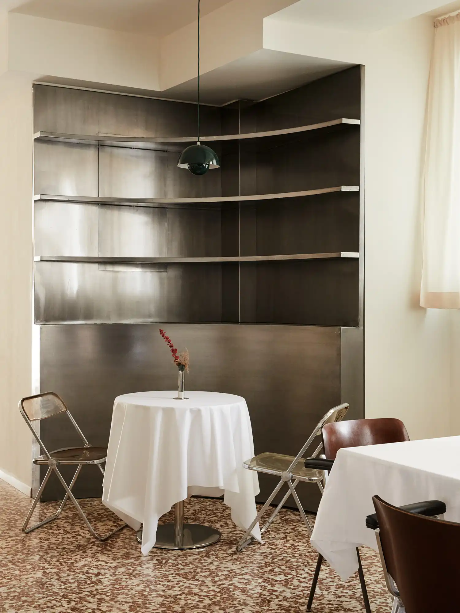 A curved, monolithic brushed metal structure serving as a wall feature at Sandì restaurant, with a single dining table set in front of it and a dark green pendant lamp hanging above.