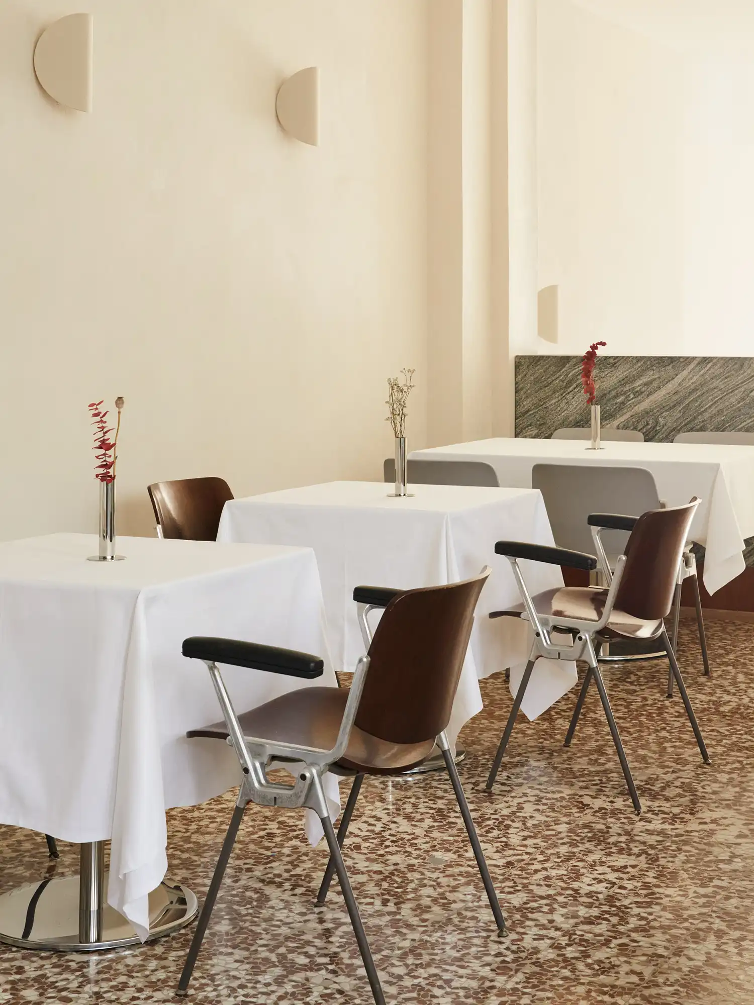 A brightly lit dining corner in Sandì restaurant, showing a white-clothed table, chairs with dark wood backrests, and a glimpse of a green marine granite base against the wall.