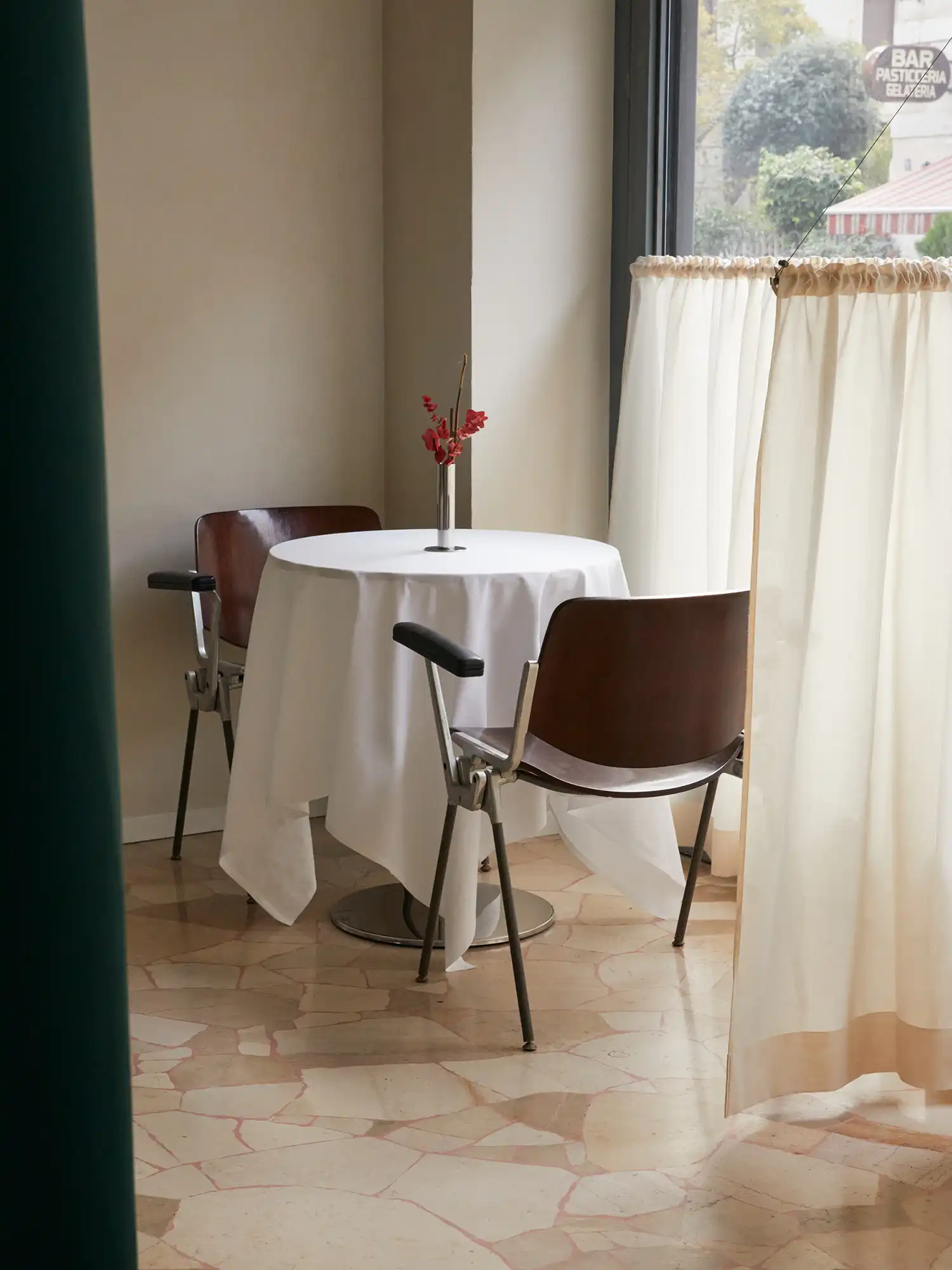 A small, intimate dining table for two in a corner of Sandì restaurant, set with a white tablecloth, a silver vase, and dark wood chairs, framed by a dark green curtain and a window with white drapes.