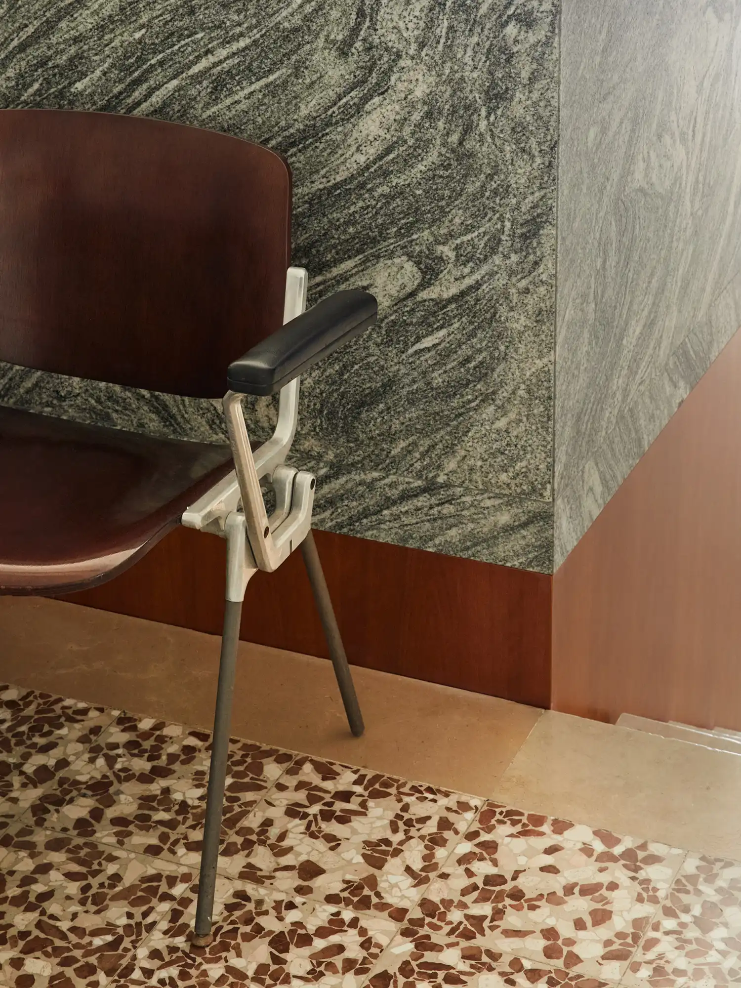 A detailed close-up of a chair with a dark wood backrest and a metallic frame placed next to a corner constructed from green marine granite panels at Sandì restaurant.