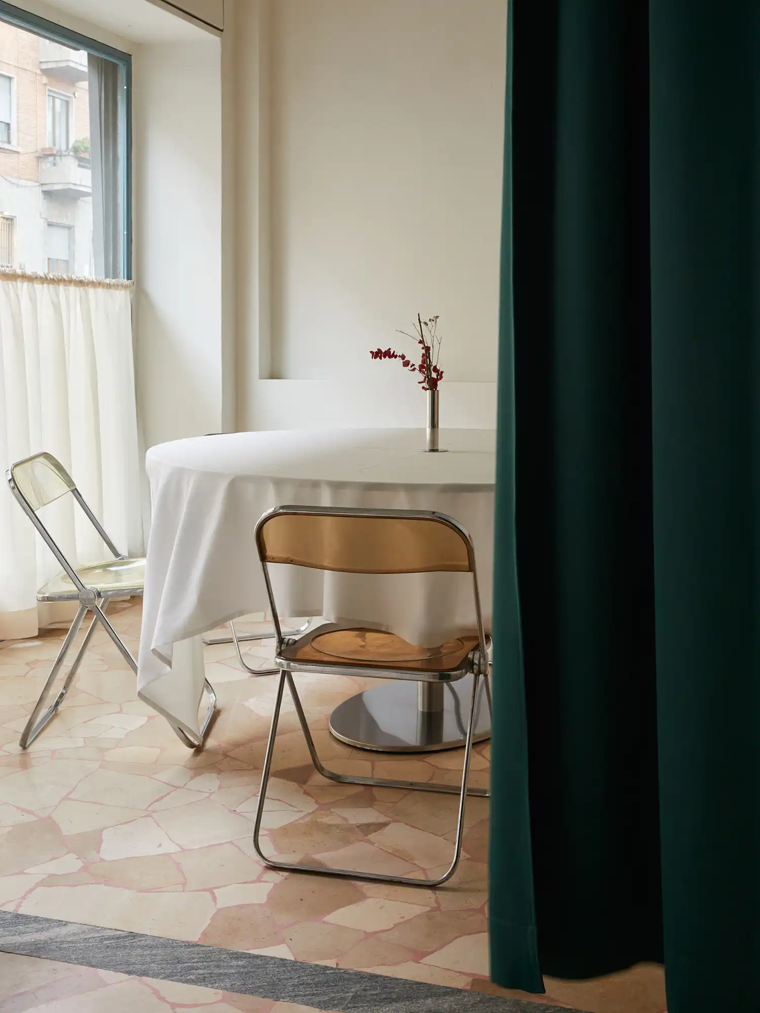A view of a round table set for dining at Sandì restaurant, positioned next to a window with white curtains and partially obscured by a dark green curtain in the foreground.