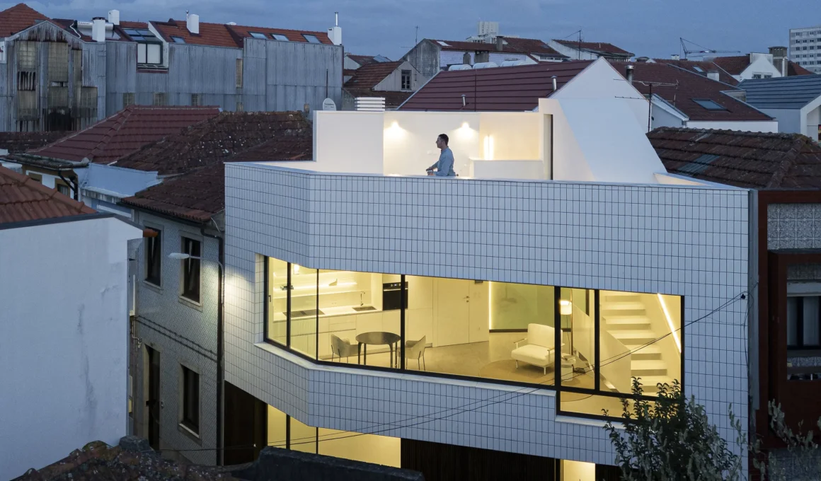 Dusk exterior view of the multi-level Beira Mar II house by Paulo Martins in Aveiro, showing the illuminated glass-walled kitchen and upper terrace with white ceramic cladding against a neighborhood of traditional tile roofs.