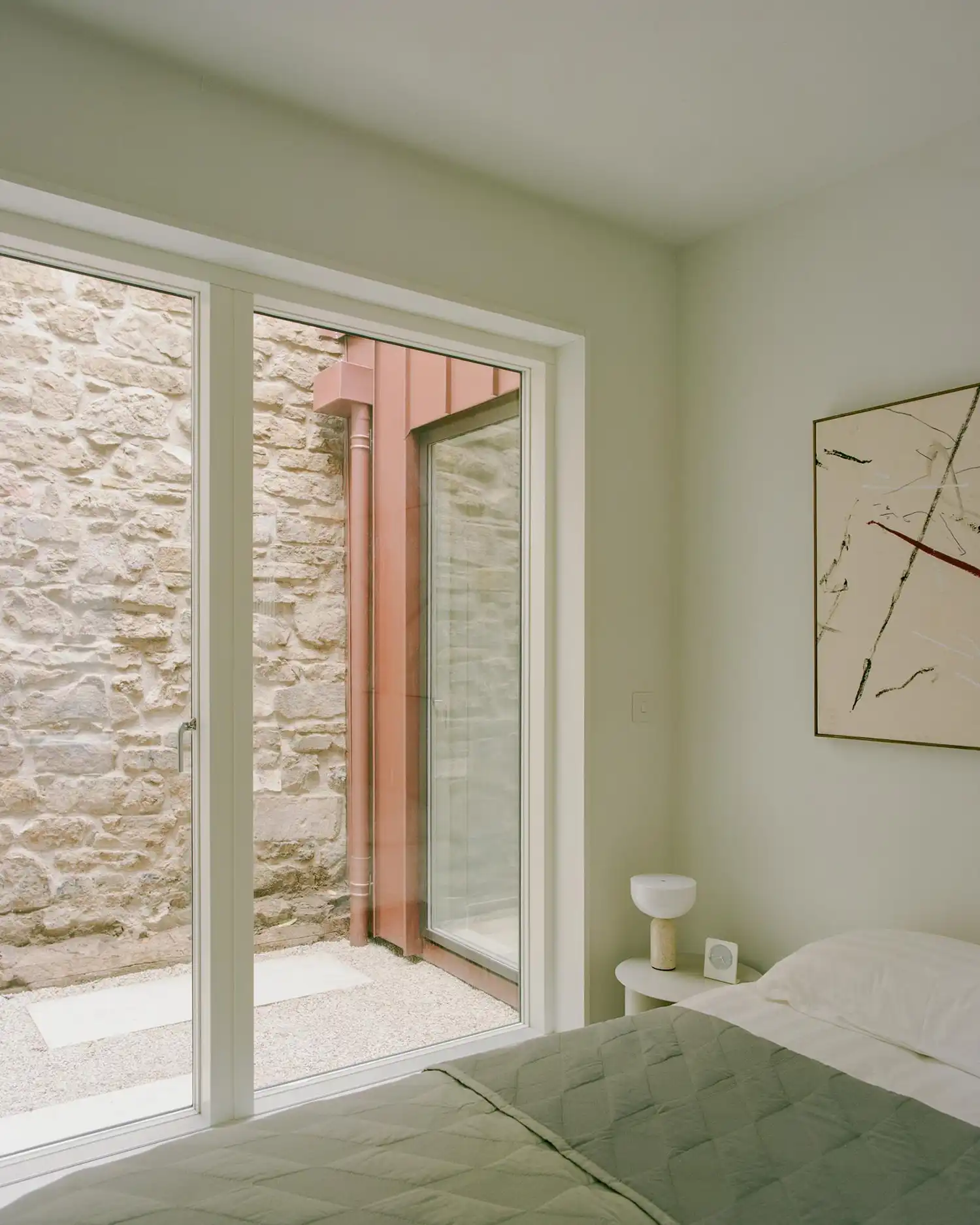 Bedroom with olive green bedding and a large window facing an old stone wall.