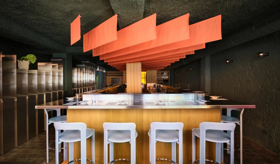 Wide interior view of MYO Madrid by Plutarco, featuring a central U-shaped bar with light wood base and stainless steel counter. Eight pastel blue stools are lined up. Over the bar, a large array of backlit peach-orange fabric panels forms an art installation. The ceiling is textured dark gray, and walls are dark stucco with metal shelving.