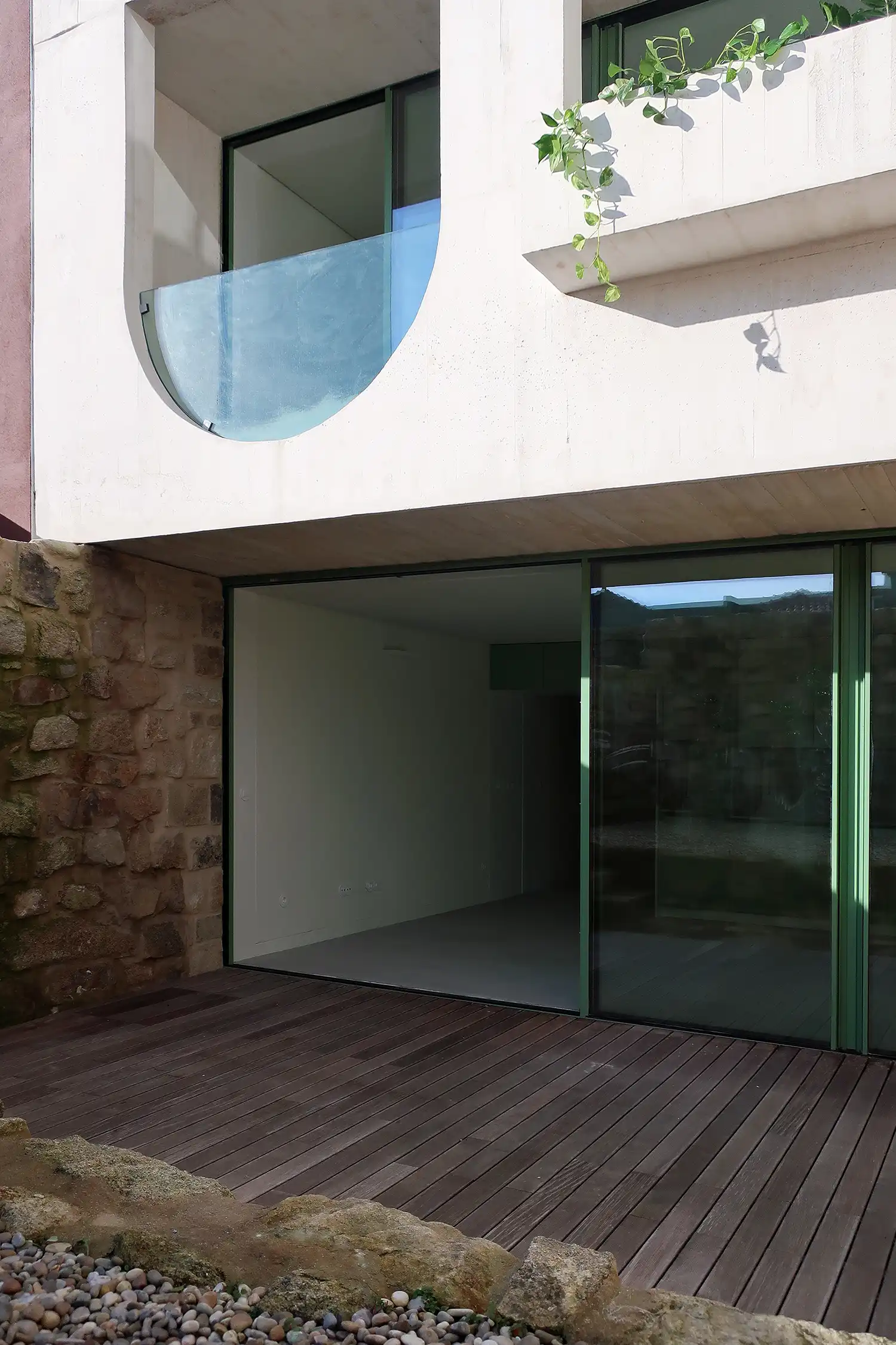 Close-up of a ground-floor terrace featuring dark wood decking and a large green-framed sliding glass door. To the left, a wall made of recycled rough stone. Above, a white concrete balcony with a semi-circular glass railing and hanging green vines.