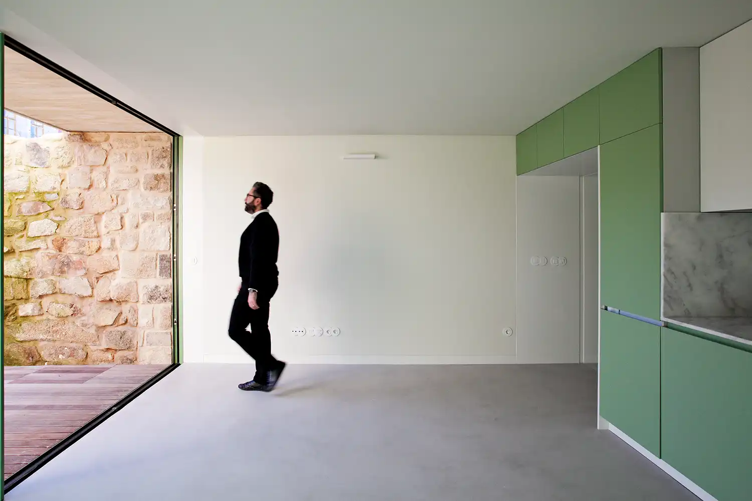 A minimalist interior with light grey concrete floors and white walls. A man in a black suit walks past a large open sliding glass door that reveals a recycled stone wall outside. To the right, integrated cabinetry in a sage green finish.