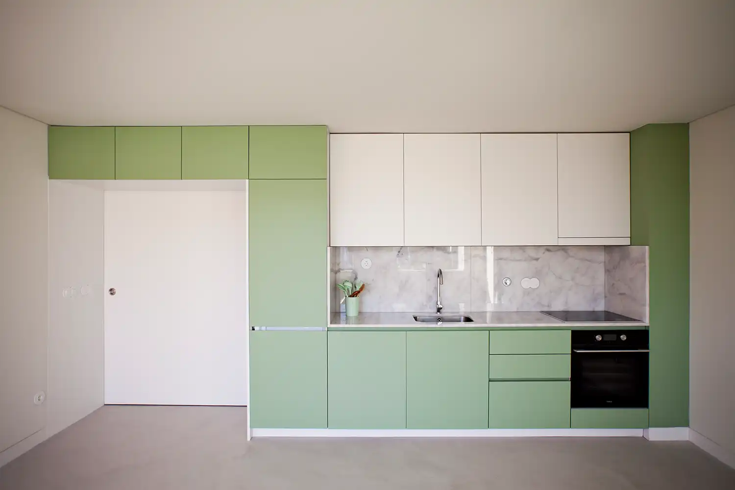 A head-on shot of a minimalist integrated kitchen. The cabinetry is a split of sage green and white. It features a marble backsplash and countertop, a stainless steel sink, and a built-in black oven. The floor is light grey.