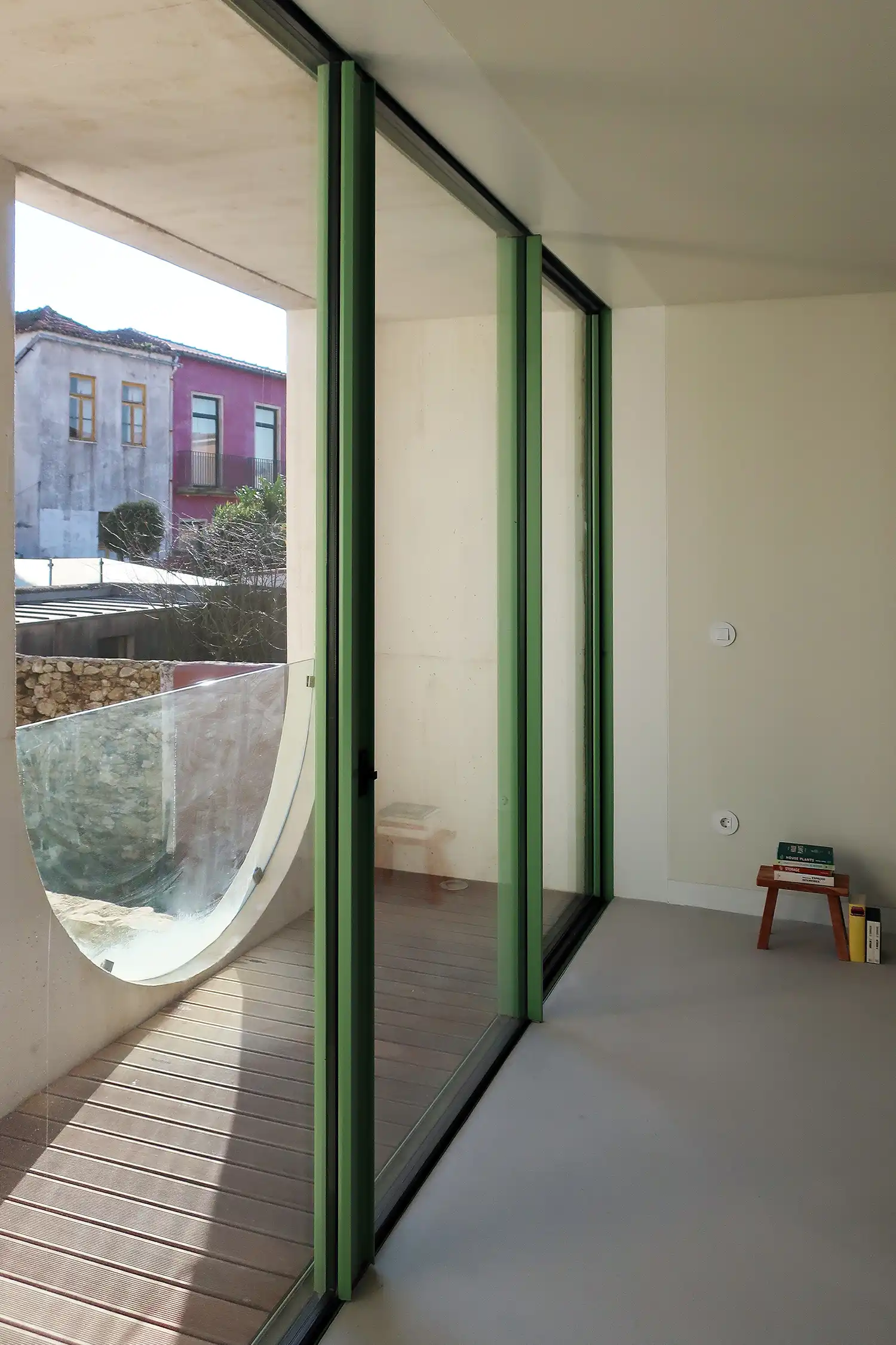 Interior view looking out from the upper floor through three green-framed glass panels. Outside is a balcony with wood decking and a semi-circular frosted glass railing. The view captures neighboring historic buildings in pink and grey.
