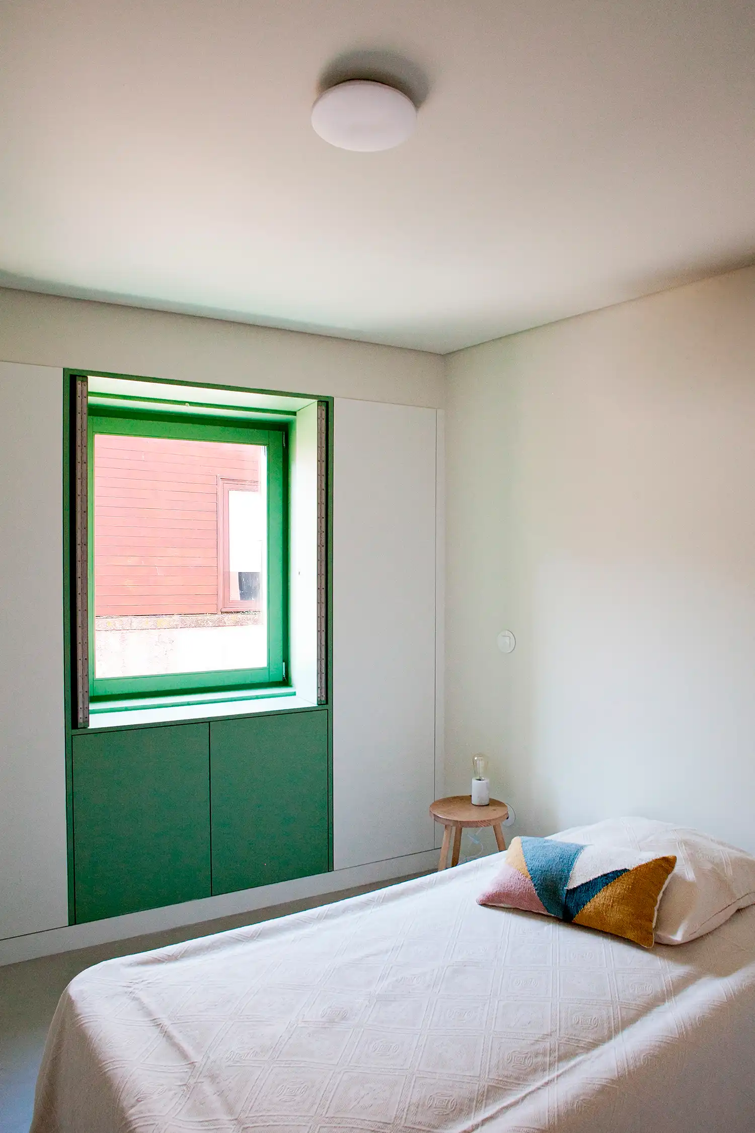 A minimalist bedroom with a single bed and a geometric-patterned pillow. A recessed window with a green frame is integrated into a wall of white and green cabinetry. A small wooden three-legged stool sits by the bed.