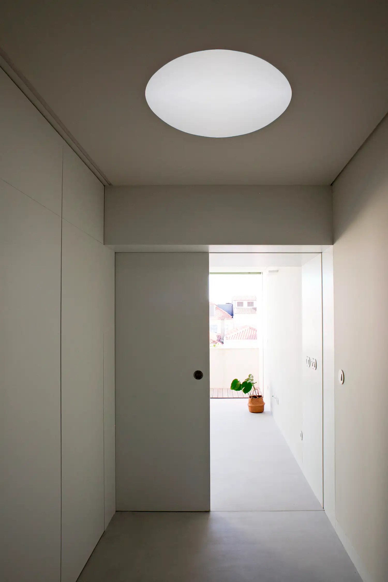 A minimal white hallway with integrated floor-to-ceiling cabinetry. A large, circular, frosted skylight in the ceiling provides soft diffused light. At the end of the hall, an open sliding door leads to a sunlit balcony with a potted plant.