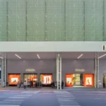 Street-level view of the Prada Fifth Avenue flagship facade in NYC, showcasing the vibrant orange window displays and the translucent green moiré scaffolding.