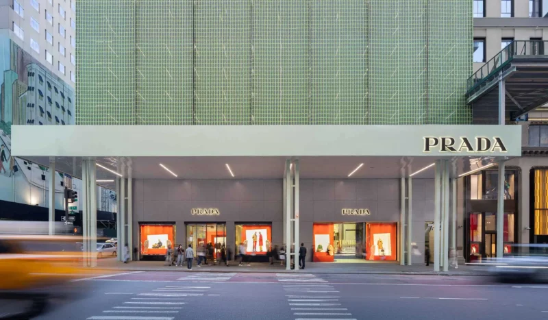 Street-level view of the Prada Fifth Avenue flagship facade in NYC, showcasing the vibrant orange window displays and the translucent green moiré scaffolding.
