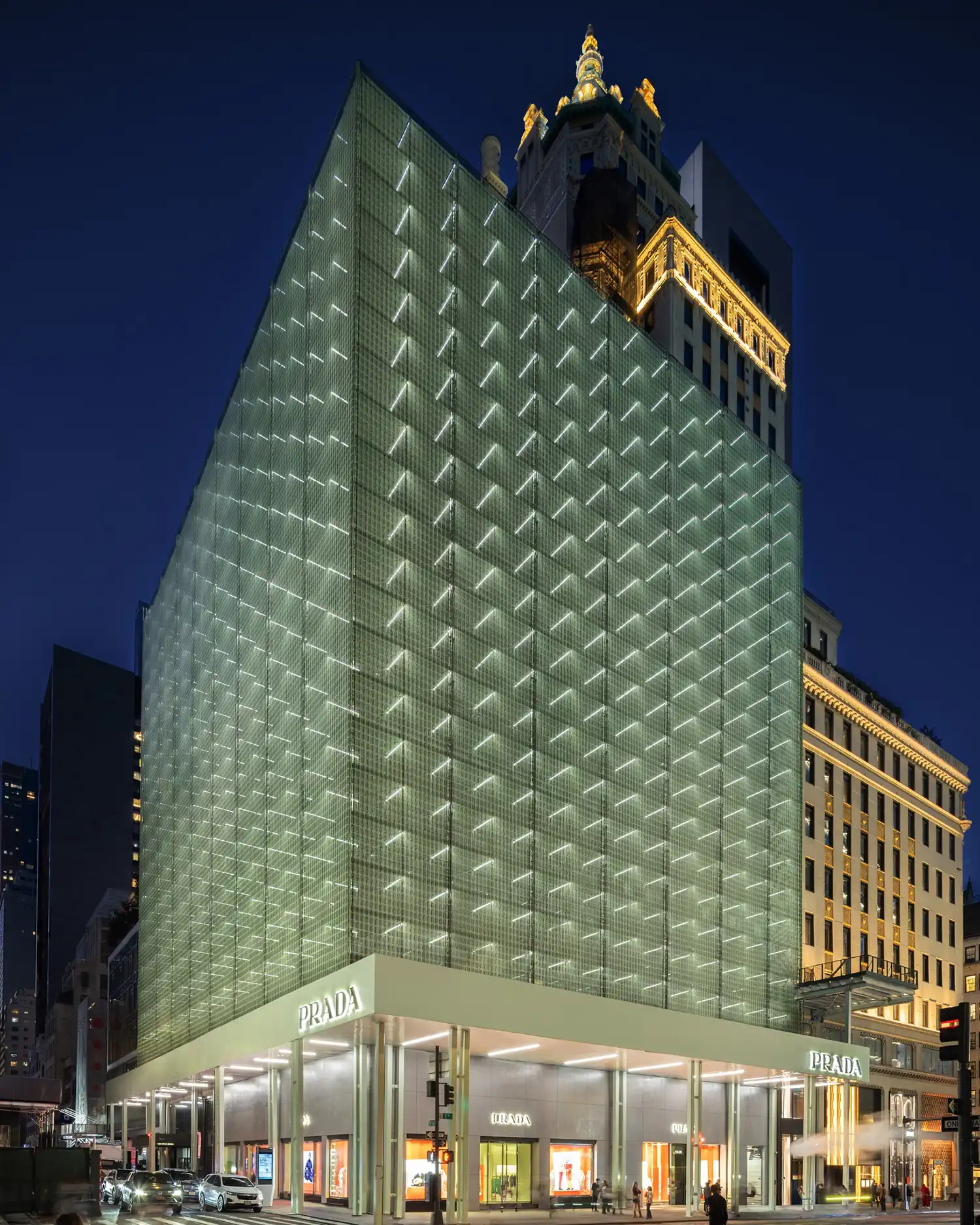 Corner perspective of the glowing Prada Fifth Avenue building at night, showing the illuminated green moiré facade contrasting with the historic neighboring architecture.