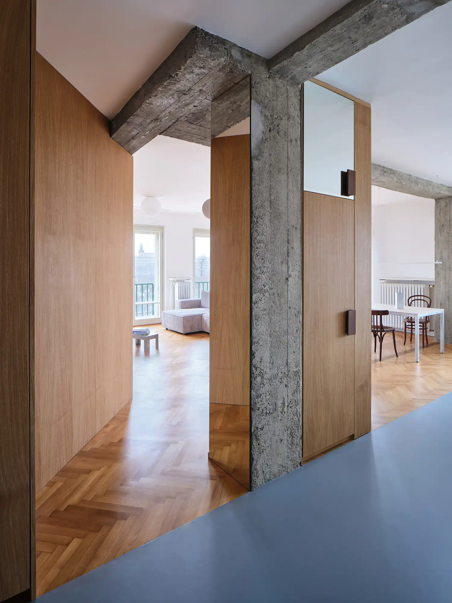 Hallway view showing oak veneer built-in wardrobes, a concrete column with a mirror, and a transition to grey polyurethane flooring.