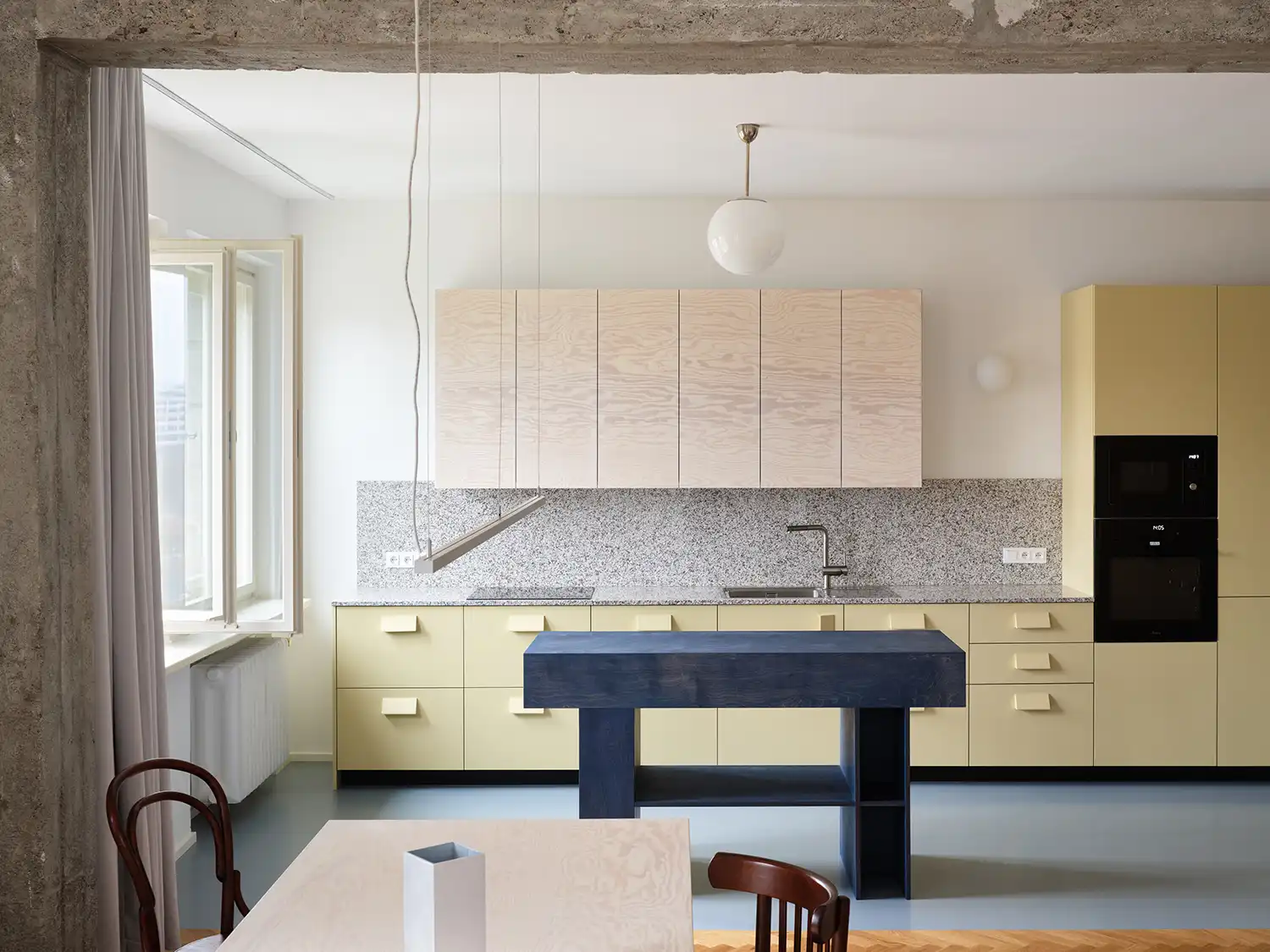 Minimalist kitchen with pale yellow lower cabinets, a blue-stained timber island, and a granite-textured splashback.