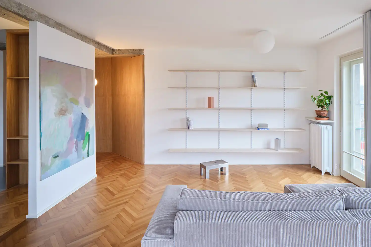 Spacious living room with a corduroy grey sofa, wall-mounted shelving, and large windows overlooking the city.