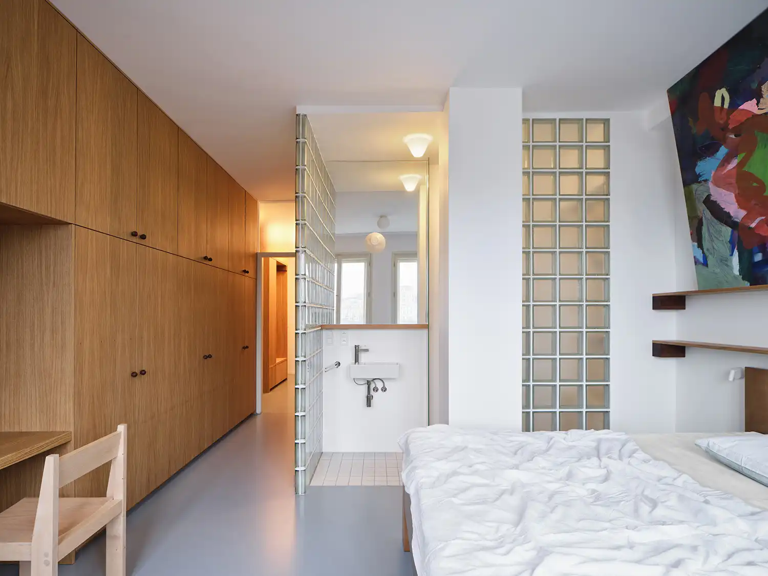 Parental bedroom featuring a wall of oak wardrobes, a white bed, and a walk-in shower separated by a glass block wall.