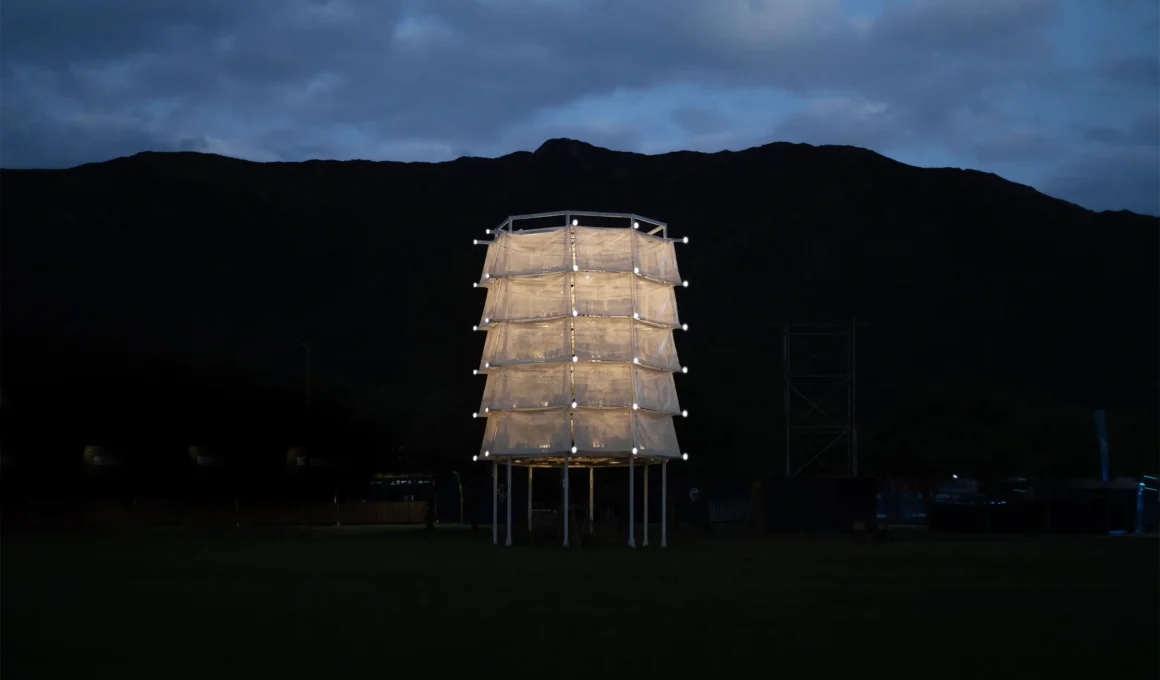 A white illuminated vertical tower installation standing in a dark field with a mountain silhouette in the background under a twilight sky.