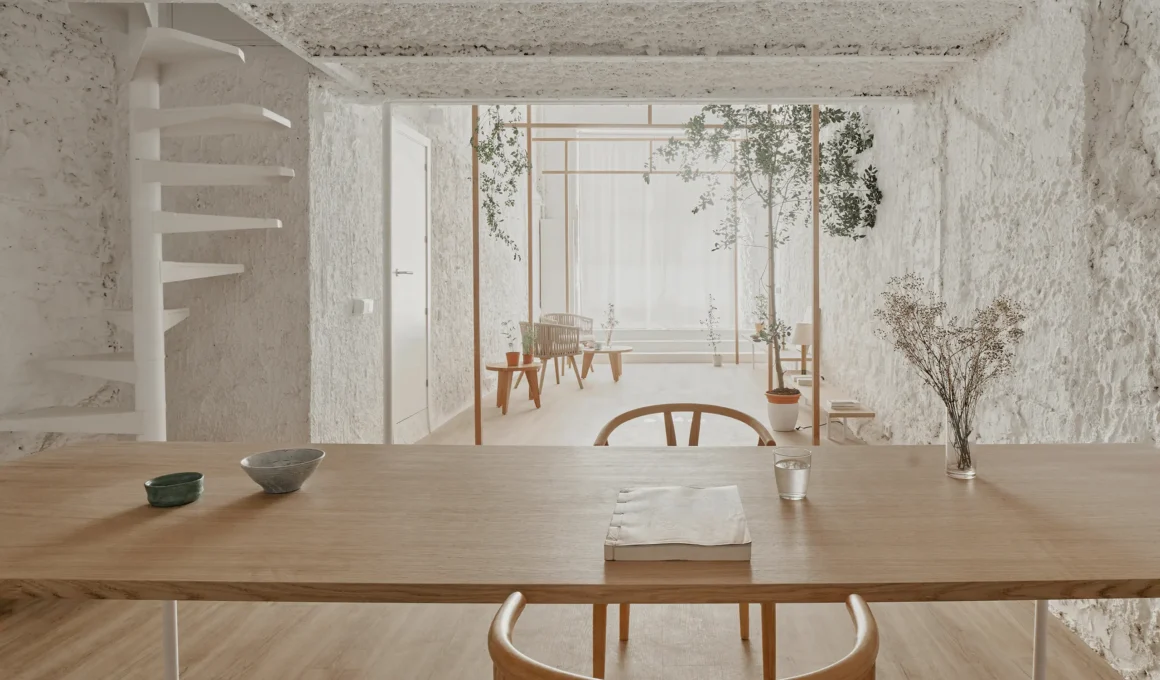 A minimalist wooden dining table in the foreground with a view of a slender ash wood torii frame and indoor trees against white textured walls.