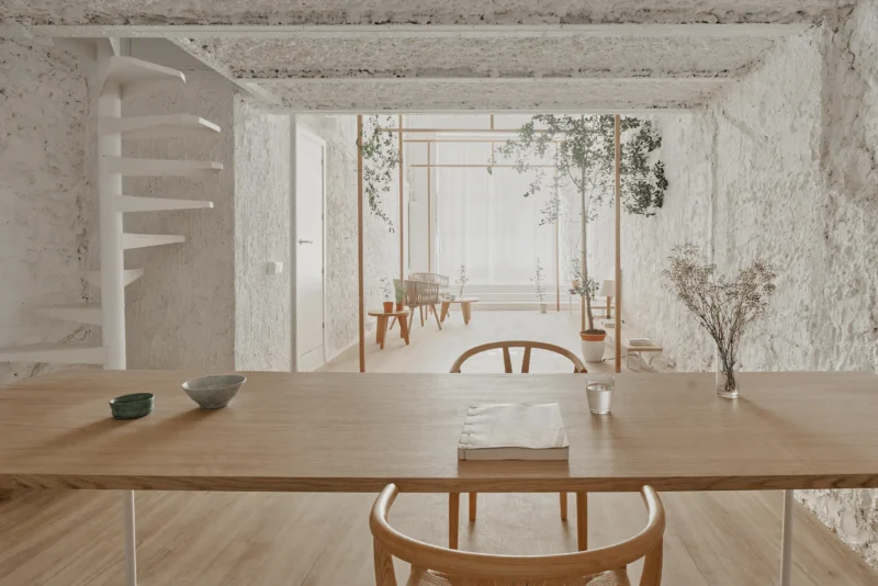 A minimalist wooden dining table in the foreground with a view of a slender ash wood torii frame and indoor trees against white textured walls.
