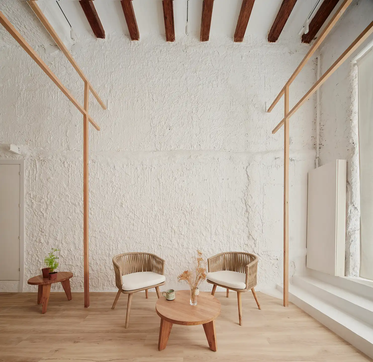 Two woven wooden armchairs and a circular low table positioned between two vertical ash wood pillars against a white wall.