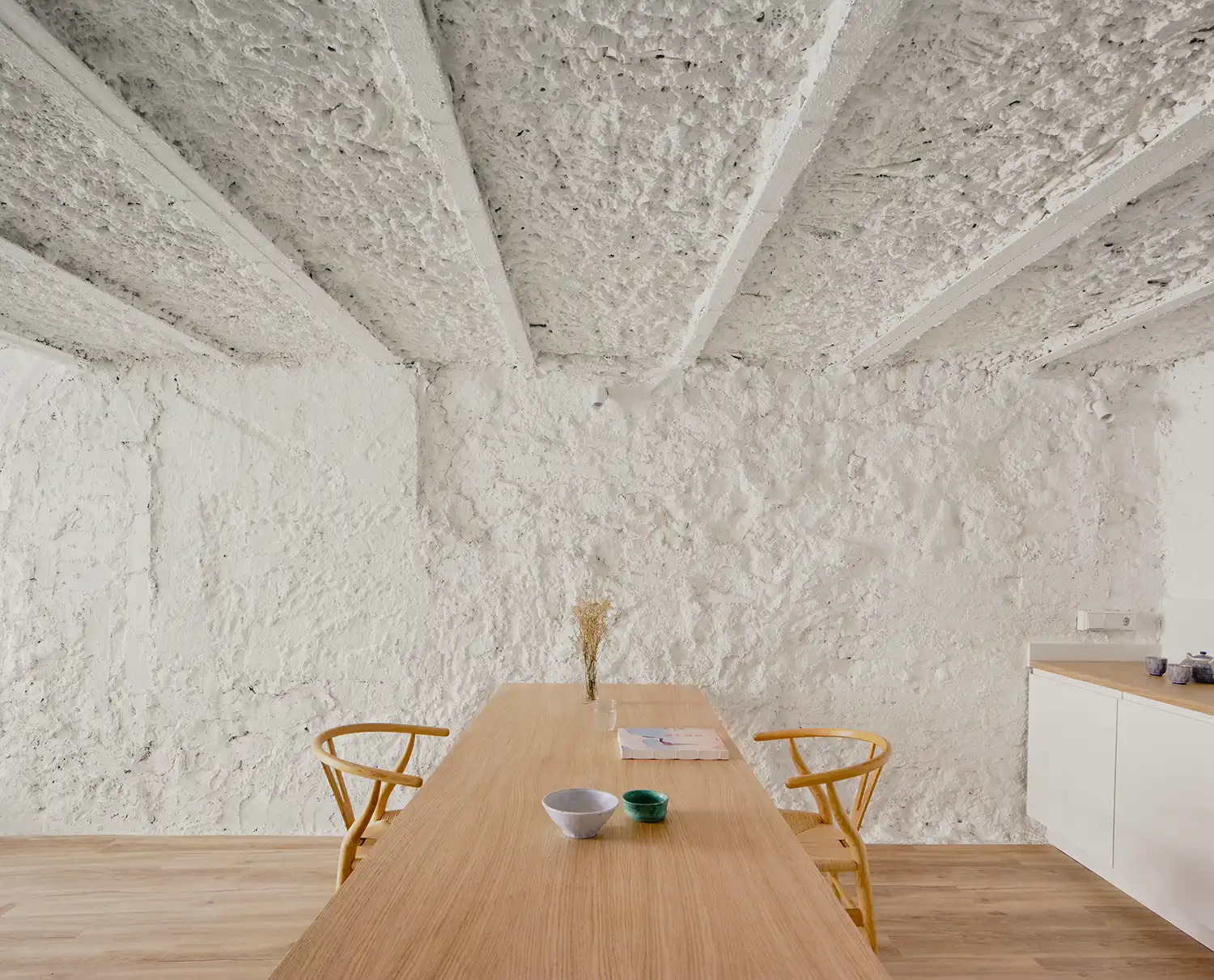 A long, slender ash wood table with two Hans Wegner-style chairs set against a rough white masonry wall.