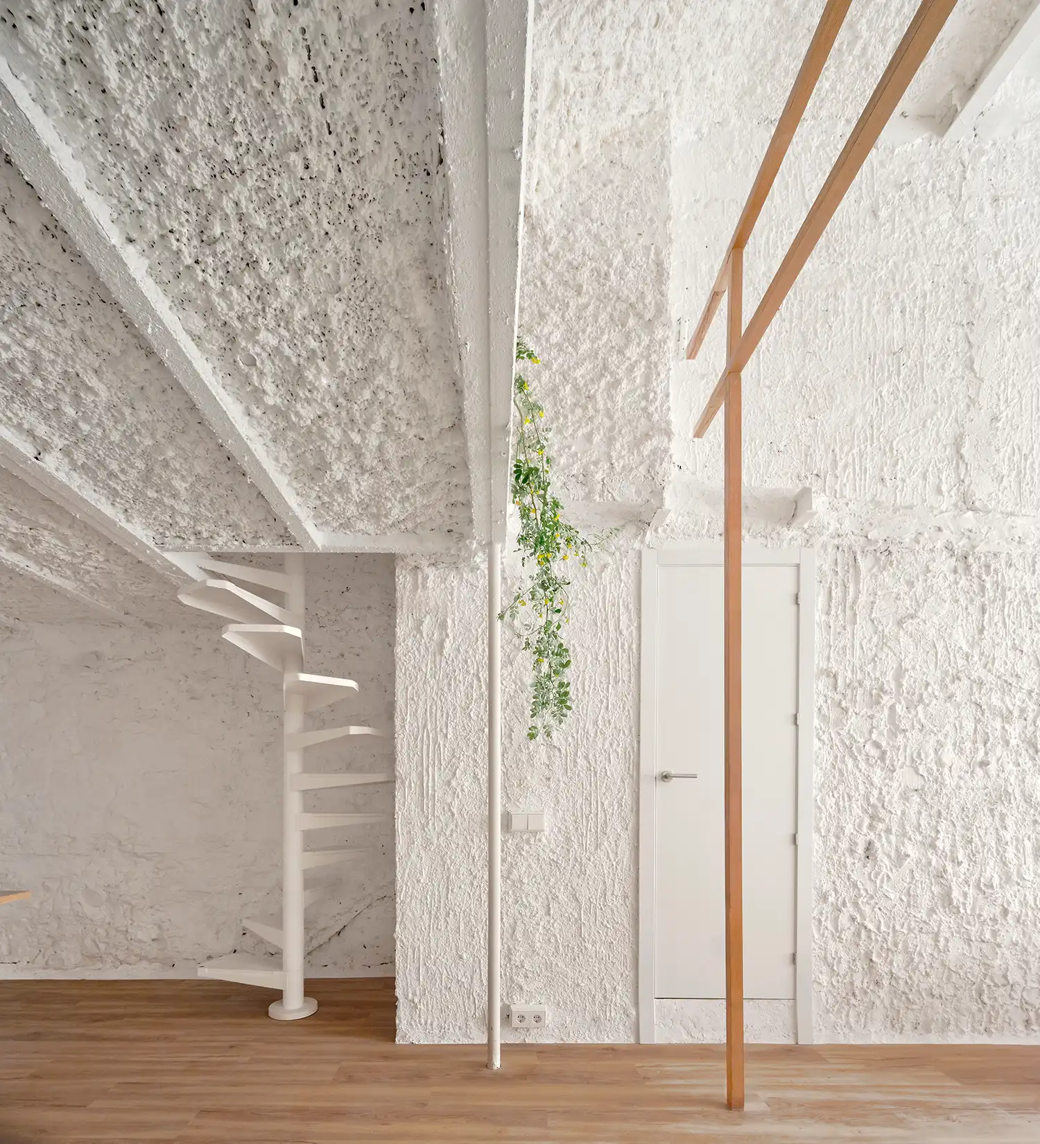 A white metal spiral staircase next to a white door and a vertical timber pillar against a heavily textured white wall.