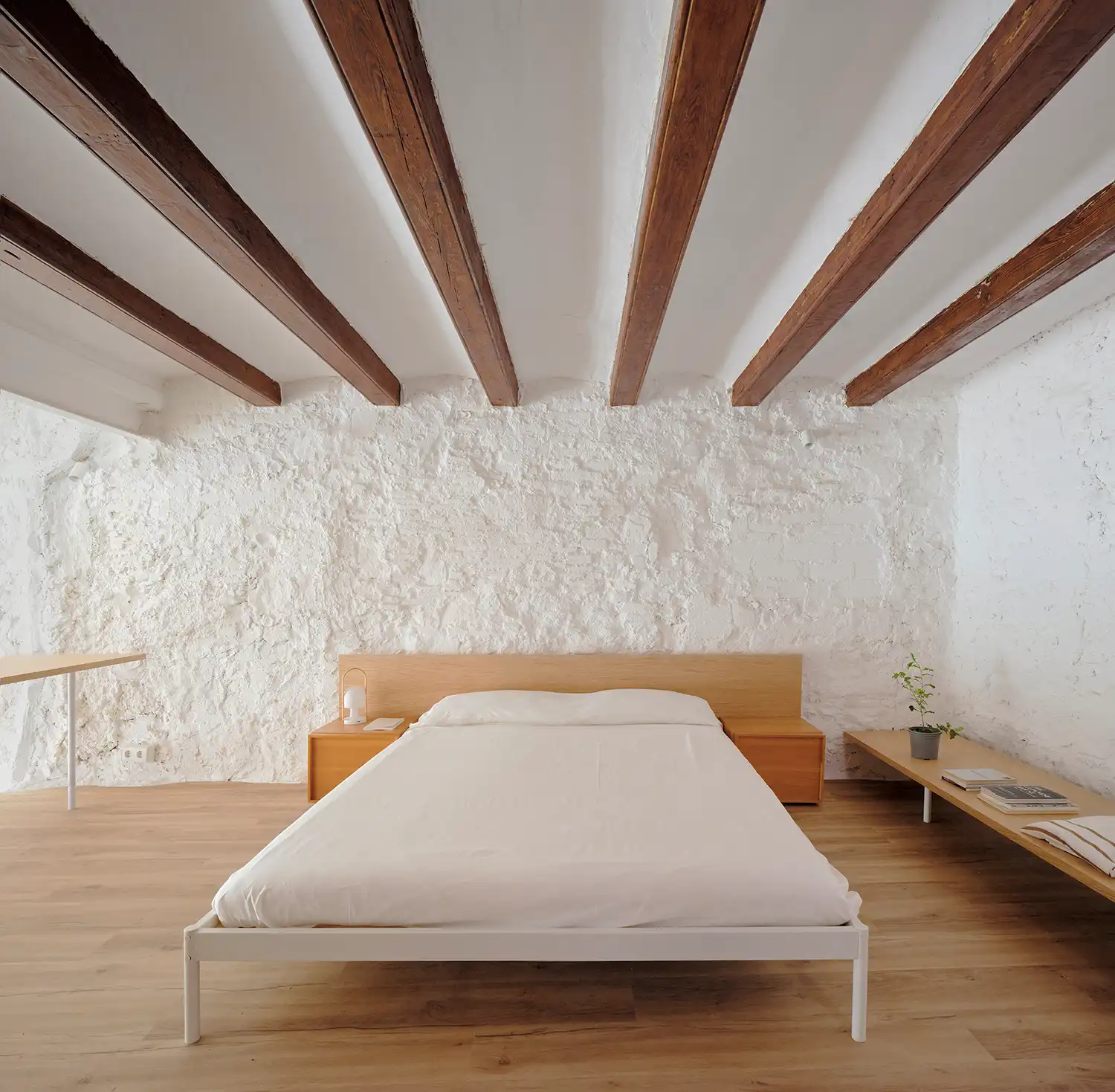 A simple white bed frame with wooden side tables and a headboard set under a low ceiling with dark wooden beams.