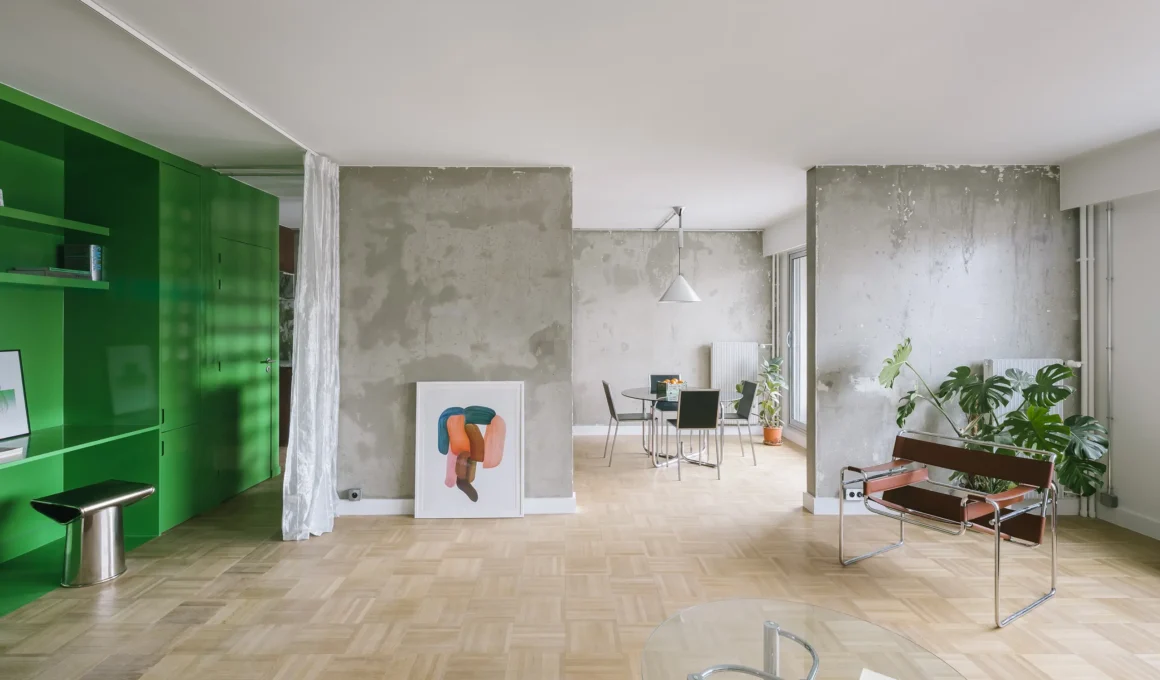 Open-plan living room in a Paris apartment featuring raw concrete walls, parquet flooring, and a glossy green structural volume.