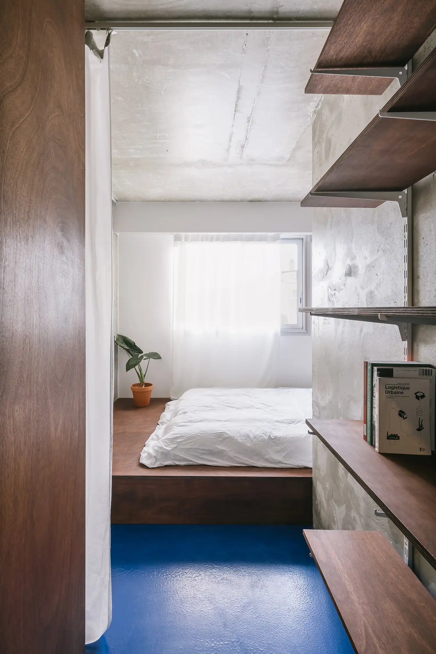 Minimalist bedroom with a platform bed, blue flooring, and simple wooden shelving on a concrete wall.