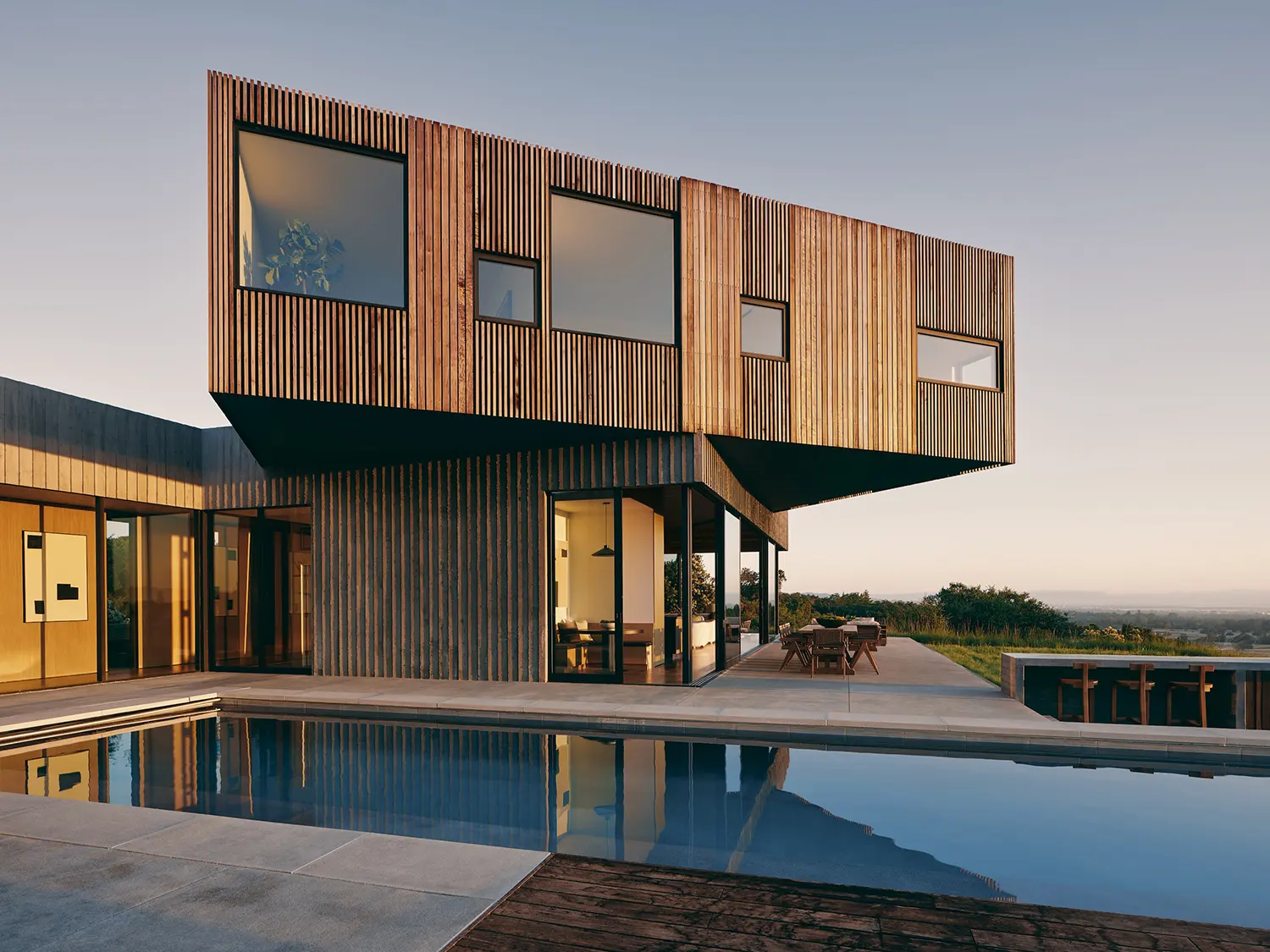 Exterior view of the house with cantilevered wood-clad volumes over an infinity pool at sunset.