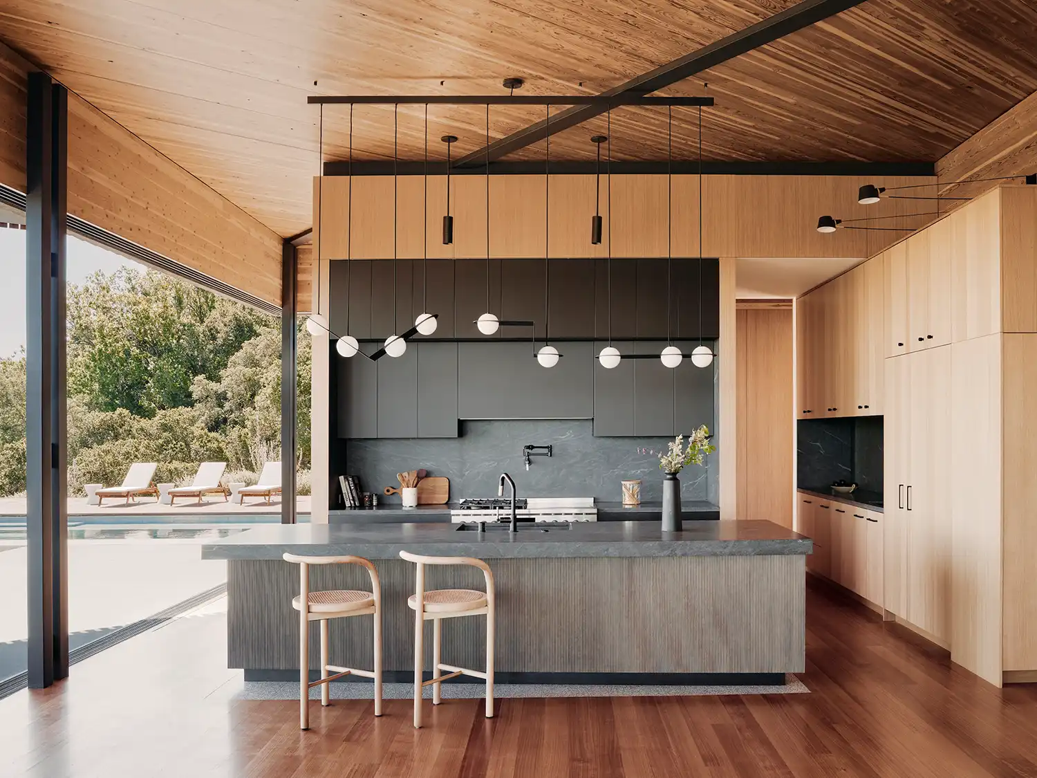 Modern kitchen with a dark stone island, light wood cabinetry, and minimalist spherical pendant lights.
