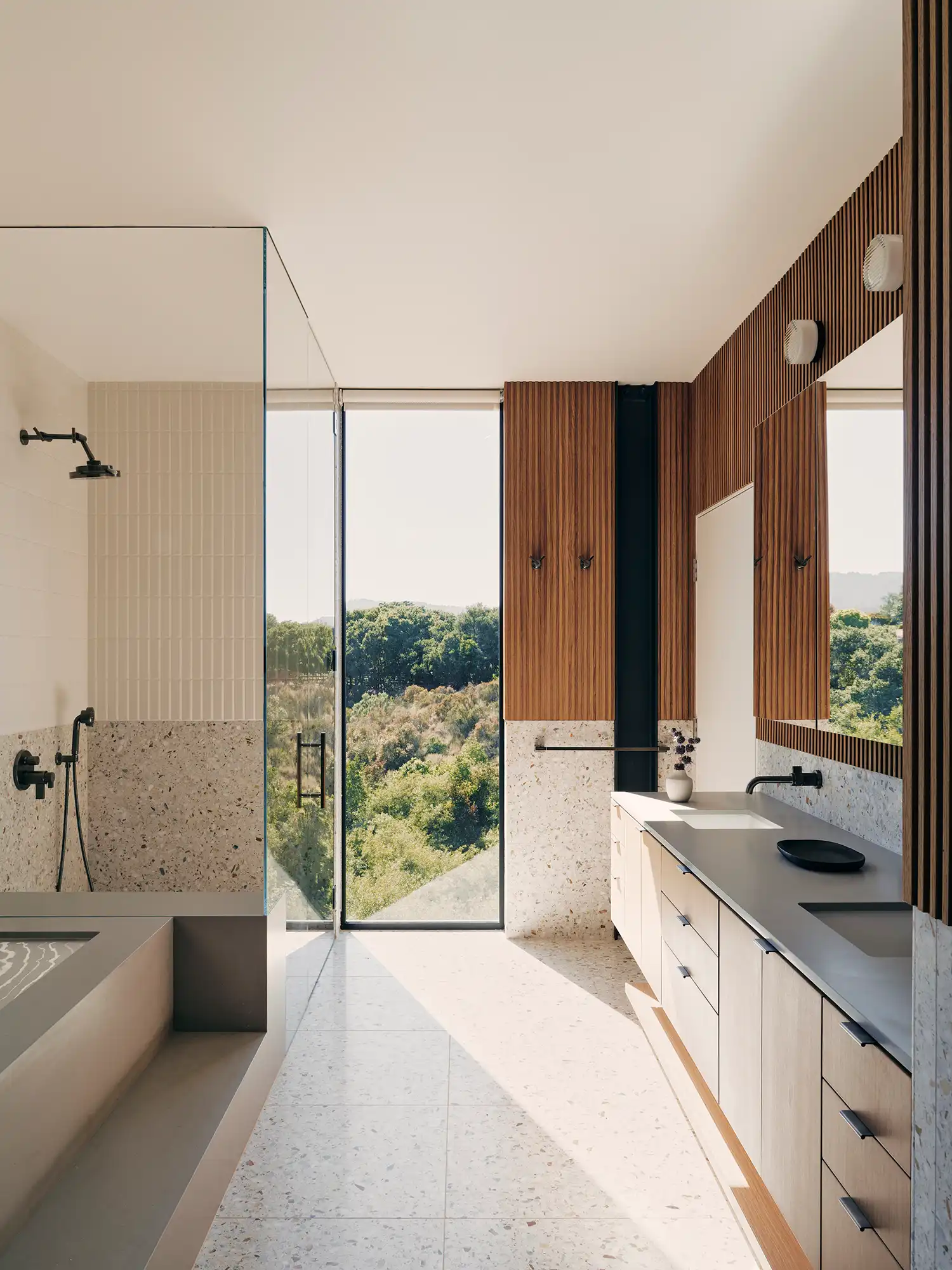 Primary bathroom with terrazzo finishes, wood-slat walls, and a large window overlooking the forest.