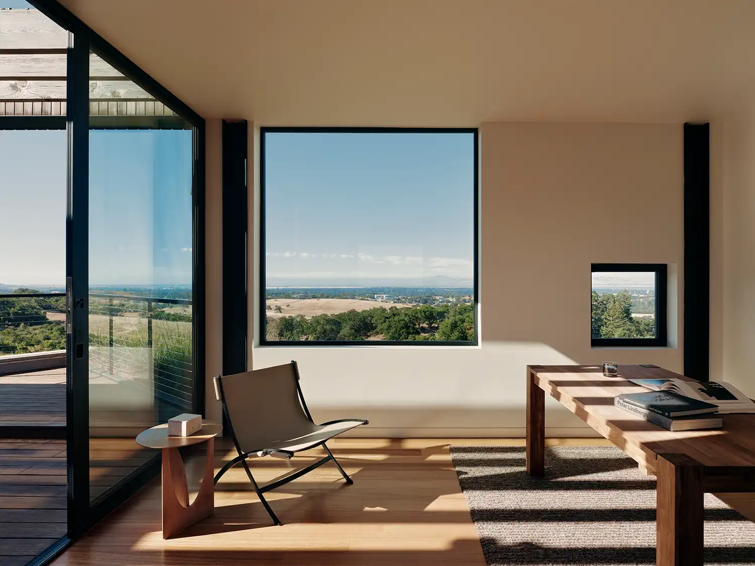 Minimalist home office with a wooden desk, leather sling chair, and large square windows overlooking the California valley.