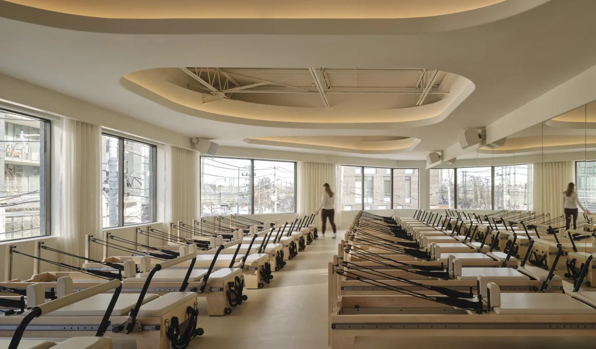A wide, daylight-filled Reformer Pilates studio in Toronto, featuring multiple rows of wood and beige Reformer machines. A person walks near the large windows that wrap the corner, with a mirror wall reflecting the space.