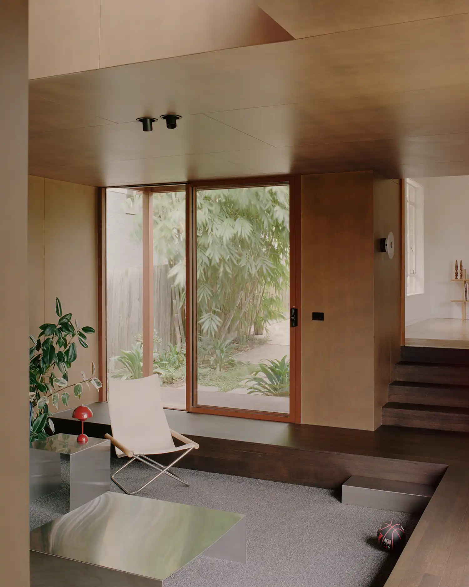 Sunken lounge area with timber steps, large sliding glass doors, and view of lush garden foliage.