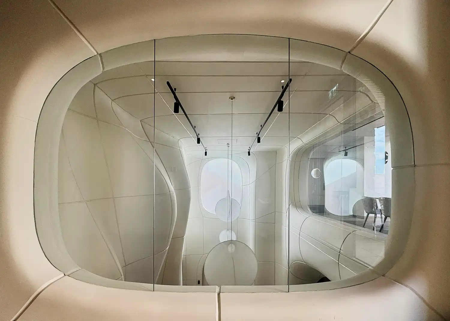 View through an oval interior window looking into a white sculptural hall with hanging lights.