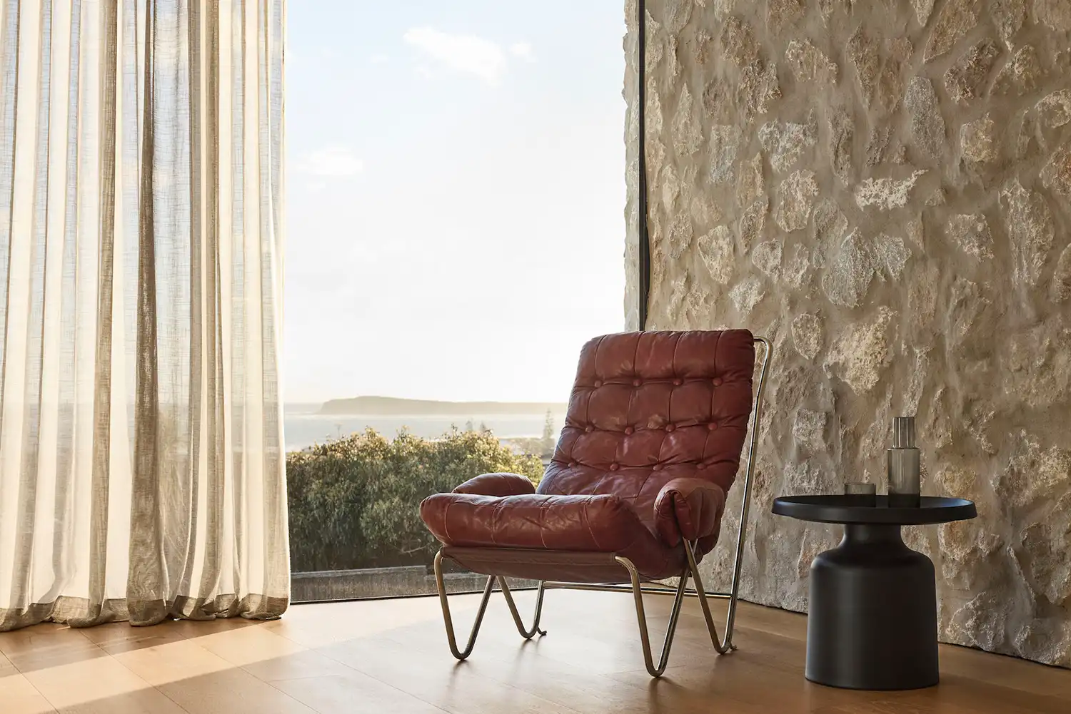 Modern interior living space with a rough limestone wall, leather lounge chair, and coastal views through sheer curtains.